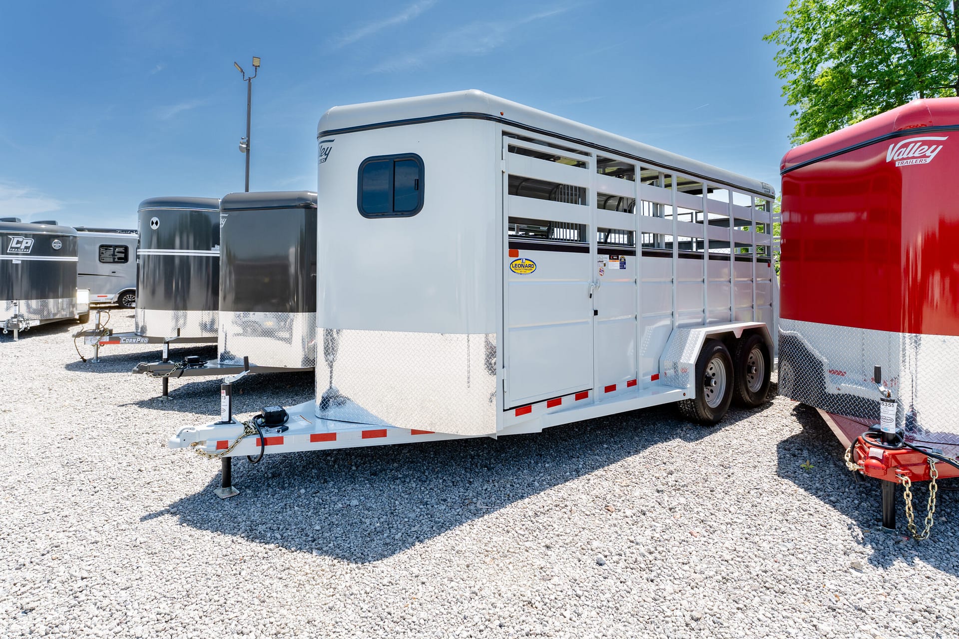White livestock trailer with open side rails parked on a gravel lot beside a red trailer, under a clear blue sky.