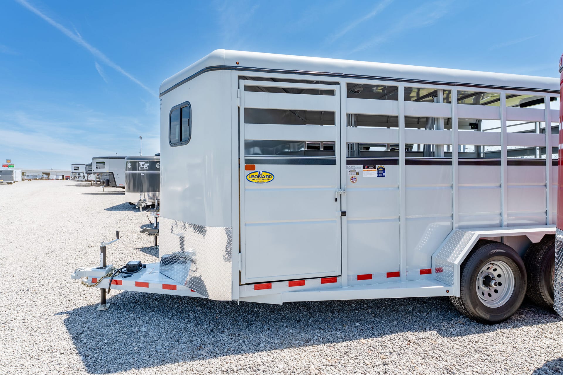 Silver livestock trailer on a gravel lot under a clear blue sky; side with ventilation slats and a small door visible at the front of the trailer's rear section.