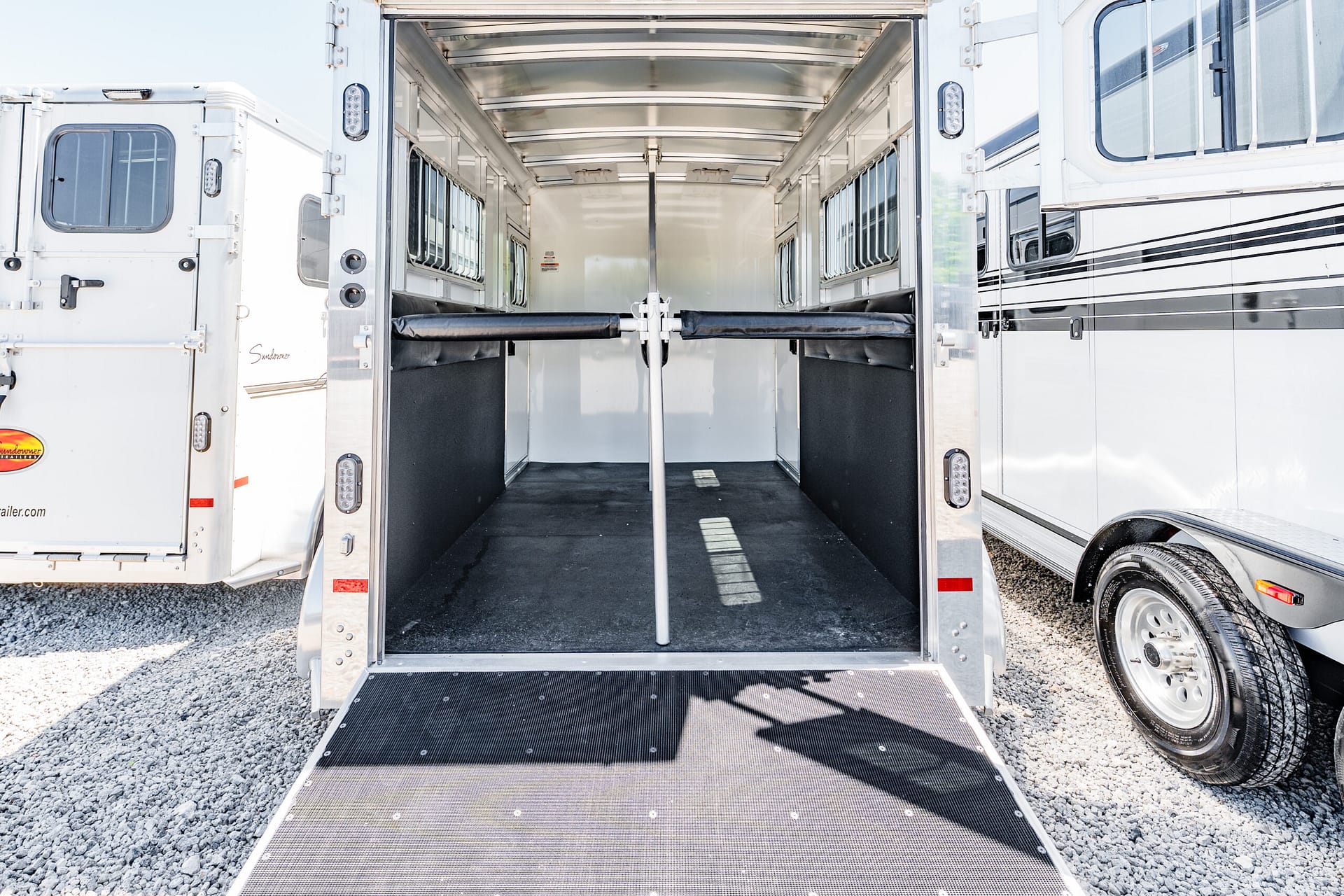 Wide view of an open horse trailer with a lowered ramp, white interior, stalls on both sides, and a middle divider bar.