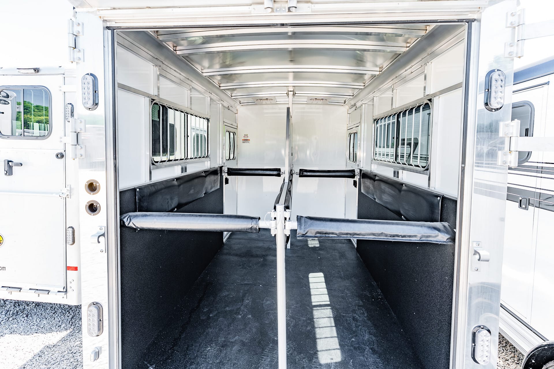 Interior of a silver livestock/horse trailer with partition rails and black stall padding, doorway open into the stalls