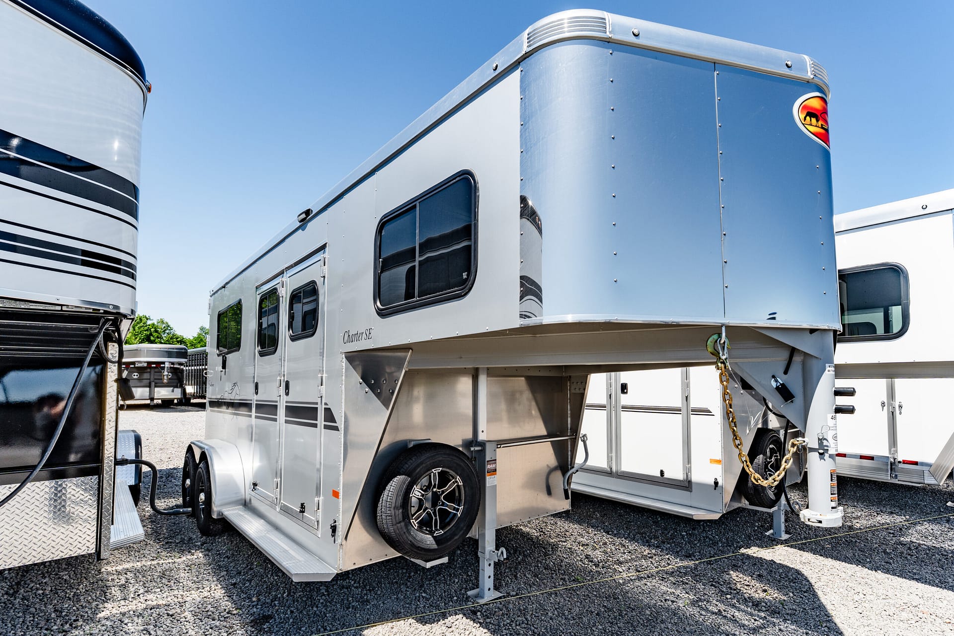 Row of aluminum horse trailers on gravel under a clear blue sky, showing windows and wheels.