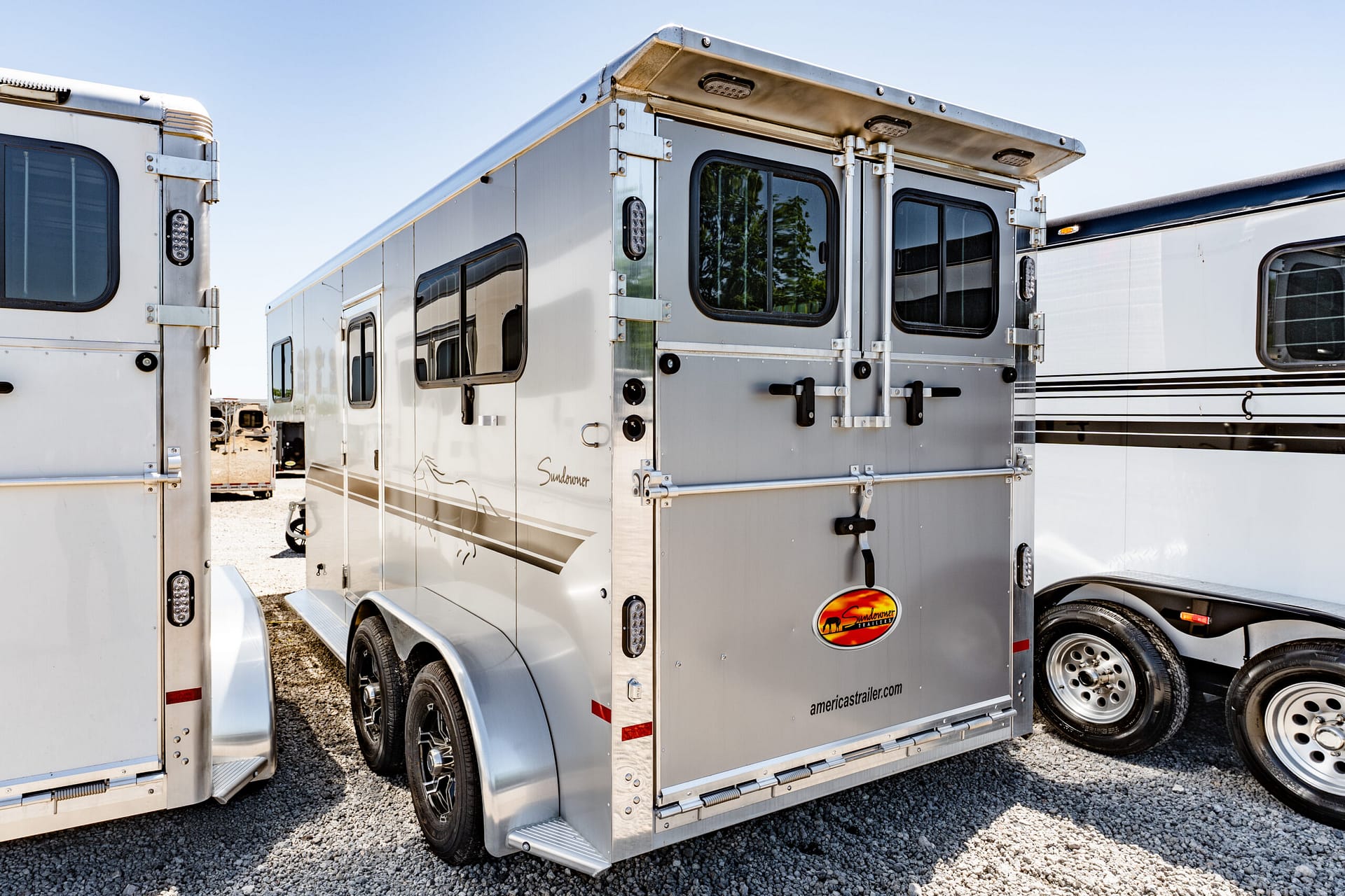 Row of silver enclosed trailers parked on a gravel lot under a blue sky, with windows and doors visible on each trailer.