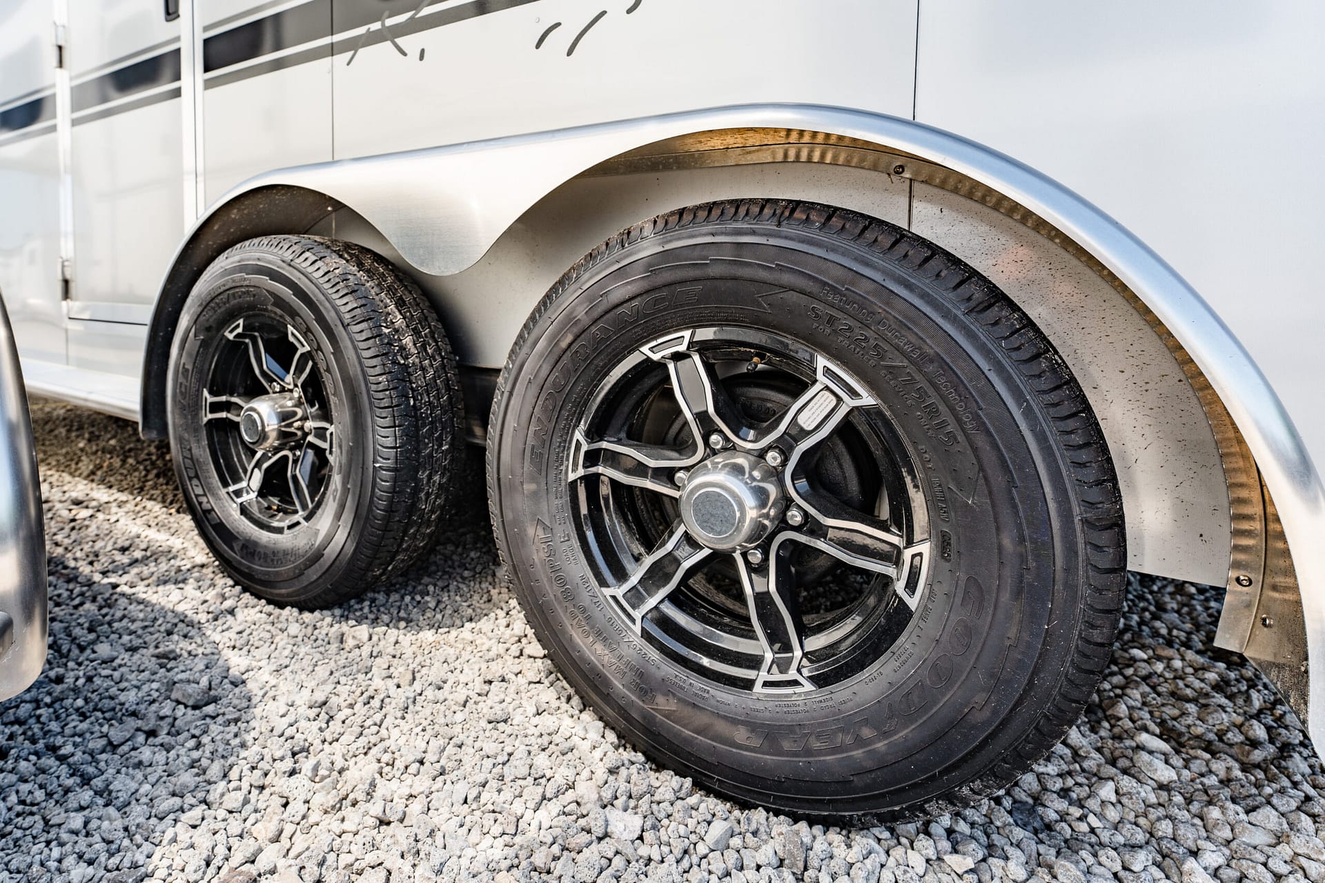Close-up of two black alloy wheels on a silver trailer with curved metal fenders, parked on gravel surface