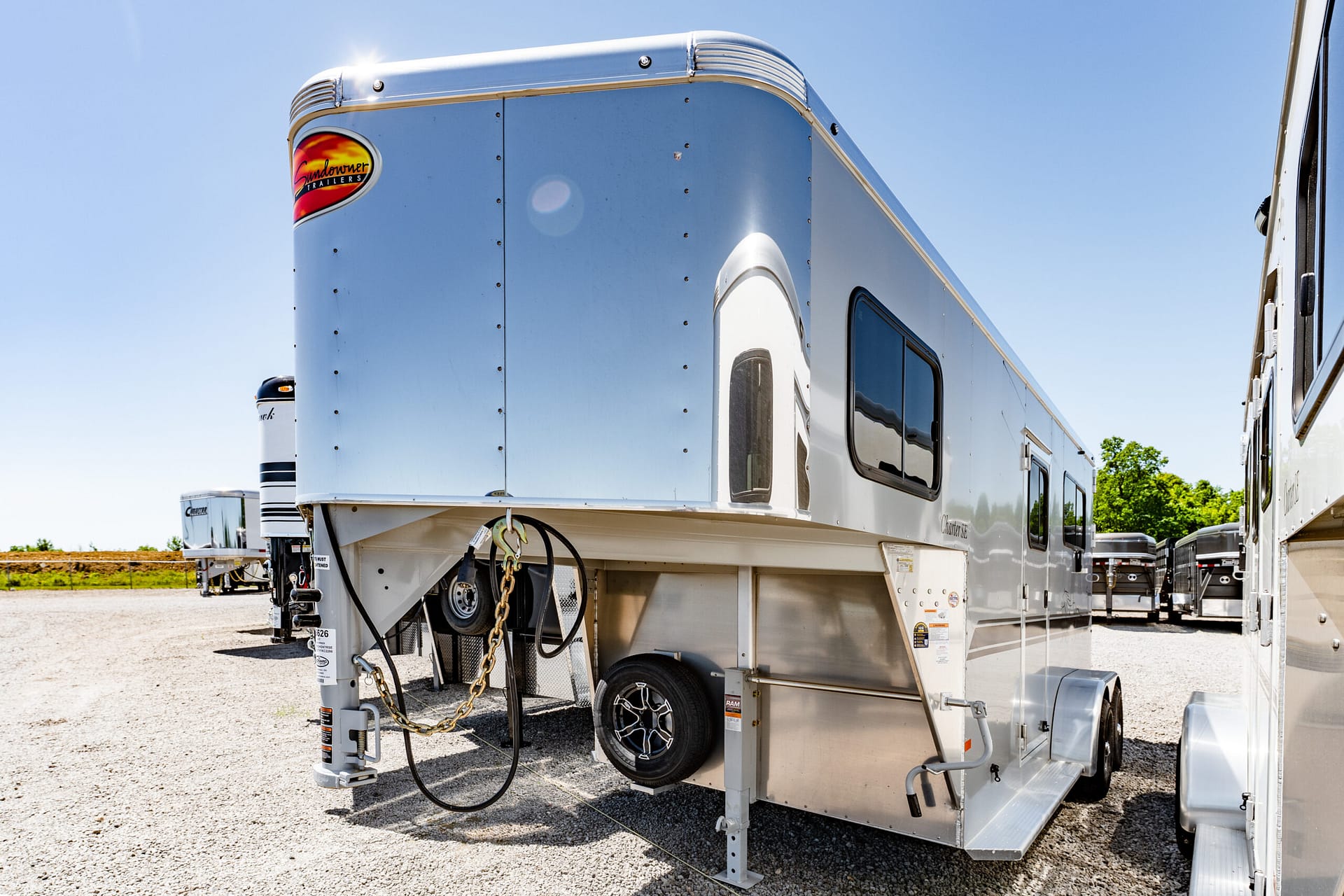 Silver travel trailer with a Sundowner Trailers logo on the front corner, parked on gravel under a clear blue sky.