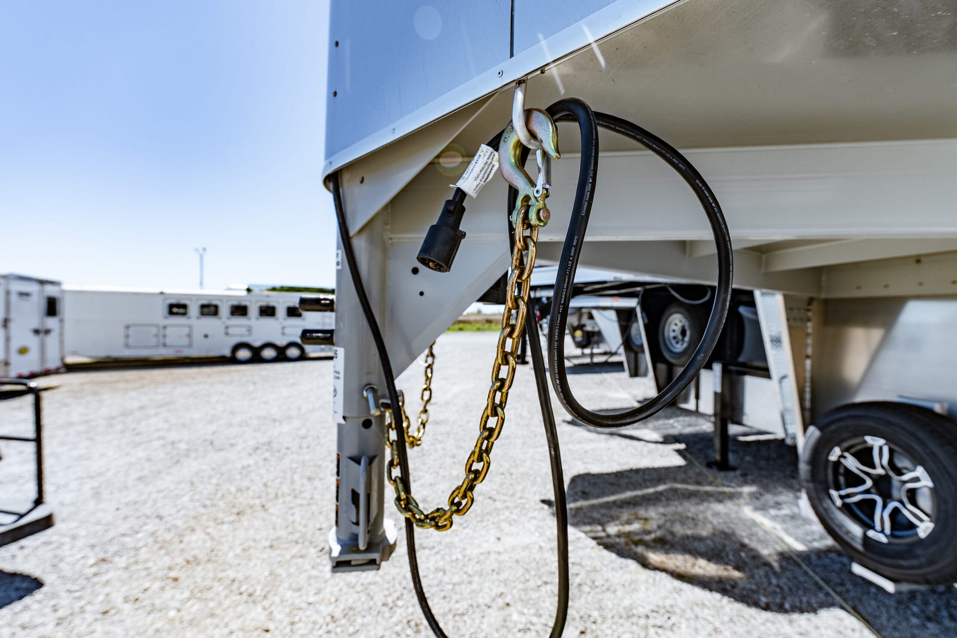 Close-up of a trailer hitch with a gold chain and black electrical cable hanging from the underside of a parked trailer, gravel lot in the background.