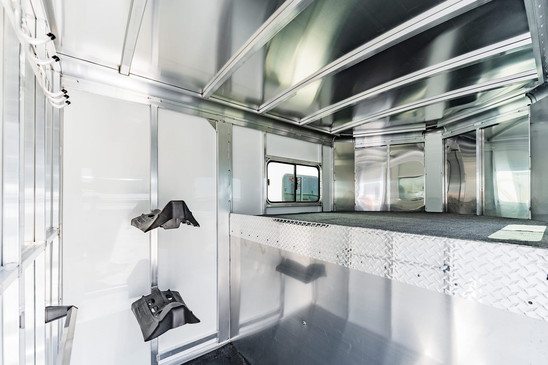 Interior of a silver aluminum trailer with metal walls, a diamond-plate floor, and black boot shells mounted on hooks along the wall near the left side.