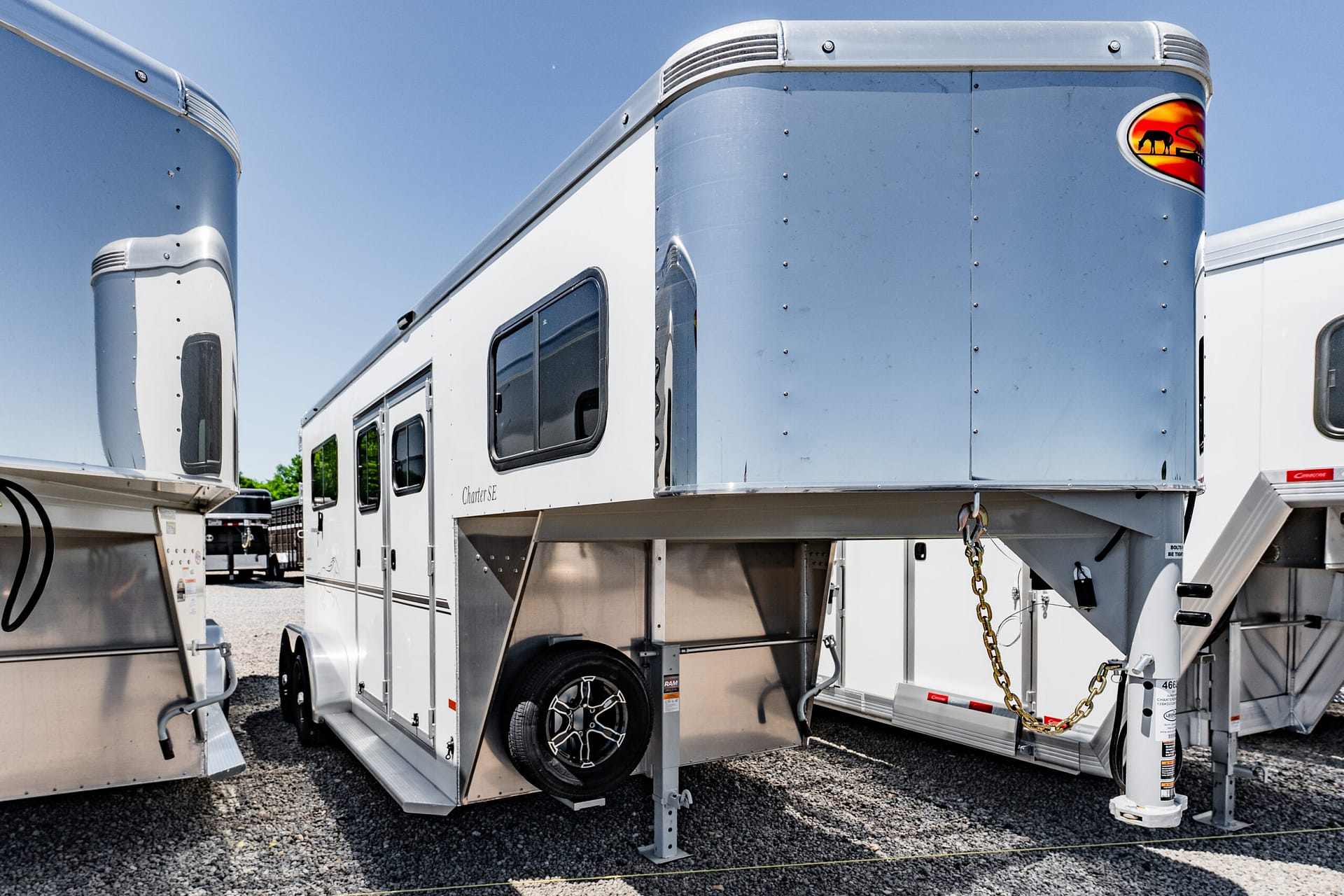 Shiny aluminum horse trailer with riveted panels and a curved front, logo on the right, parked on gravel under a blue sky