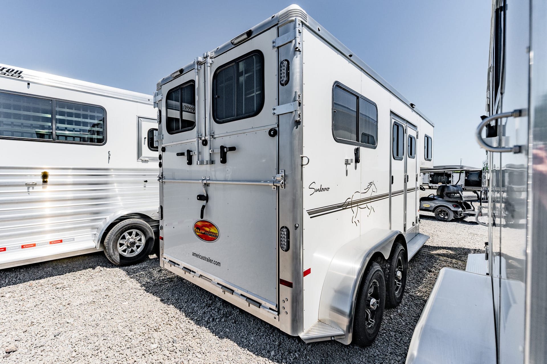 White horse trailer with two rear doors and windows parked in a gravel lot beside other trailers under a clear blue sky.