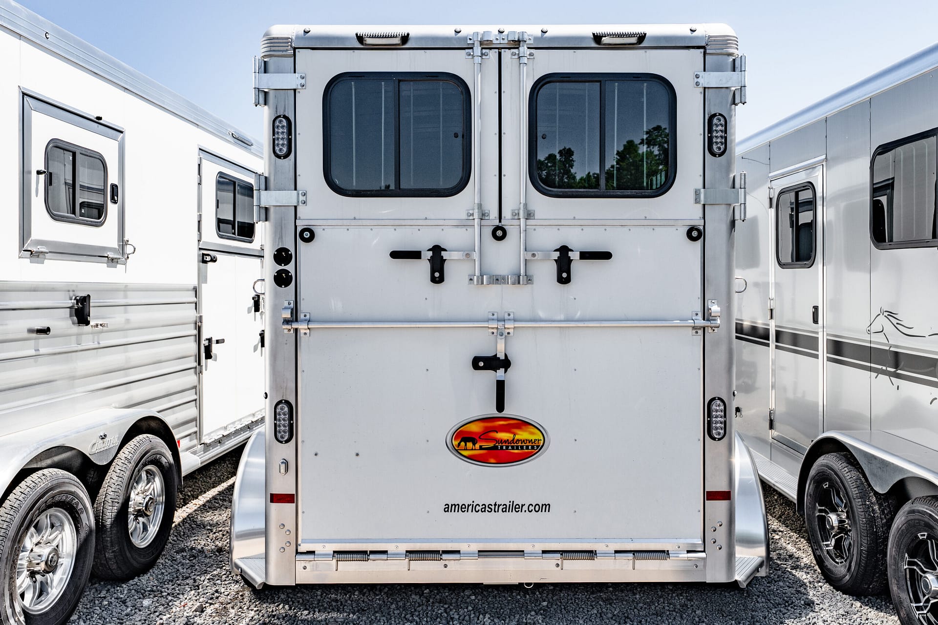 Rear of a silver horse trailer with dual doors, windows, and a Sundowner Trailers logo on a gravel lot pitured between other trailers.