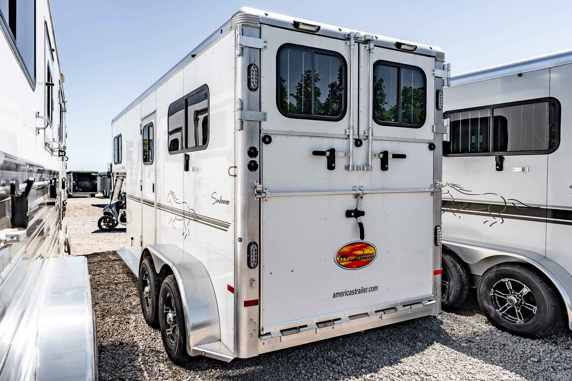 Rear view of a white horse trailer with two large back doors and windows, parked among other trailers on a gravel lot, under a clear blue sky.