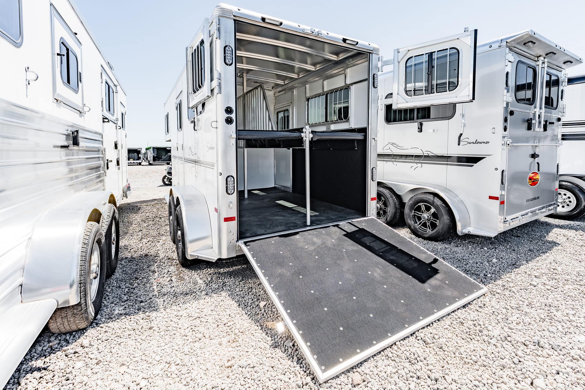 Open horse trailer with a lowered ramp, showing interior stalls and partition on a gravel lot with other trailers nearby.