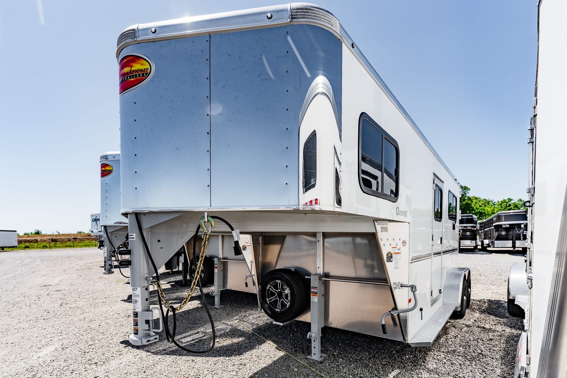 Silver fifth-wheel trailer on its landing gear in a gravel lot, with hanging safety chains and a spare wheel visible at the front.