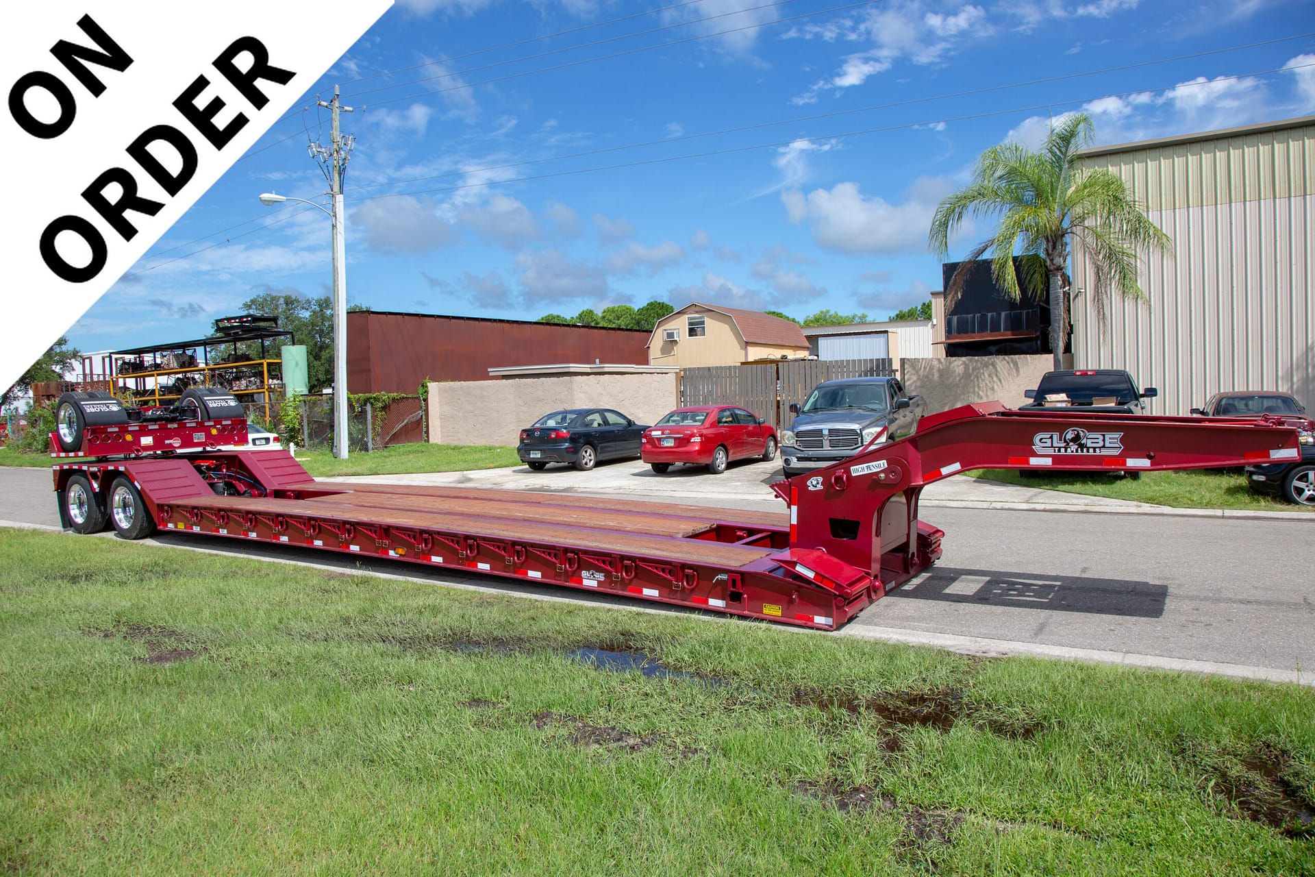 Industrial parking lot scene with a large red lowboy trailer in the foreground, beige buildings, parked cars, a palm tree, and a diagonal white banner with black text along the top-left edge (text partially visible).
