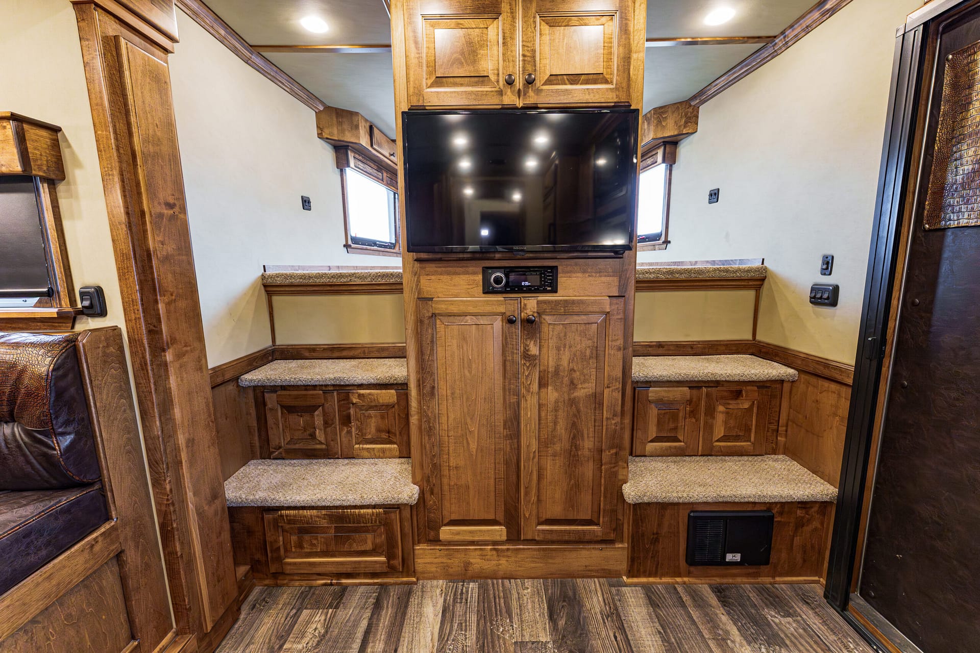Wood-paneled RV living area with a central entertainment unit, large TV mounted in the middle, cozy carpeted steps, and cabinets above and below.