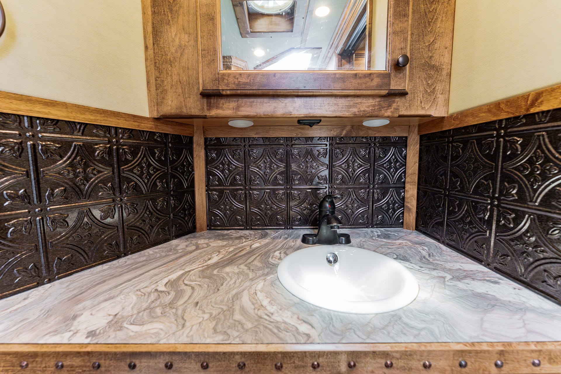 Rustic bathroom vanity with a marble countertop, dark wood frame mirror, and ornate tin tile walls with riveted trim visible behind the sink.