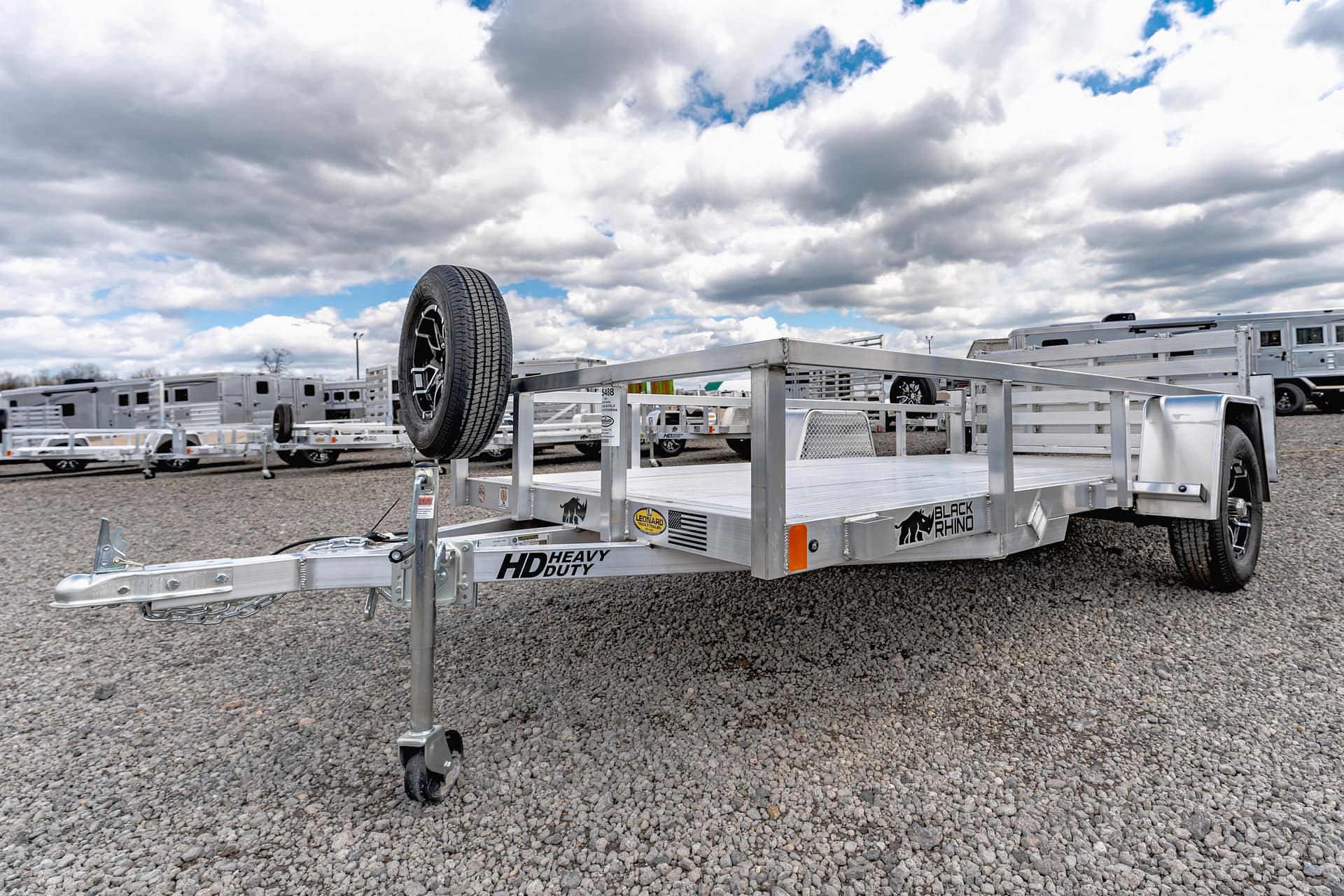Trailer with a spare tire mounted on its frame in a gravel lot, with multiple aluminum trailers in the background under a cloudy sky