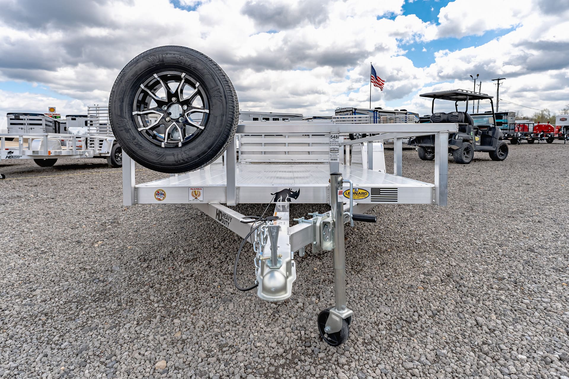 Aluminum trailer with a mounted spare tire in the foreground on a gravel lot; other trailers and an American flag in the background.