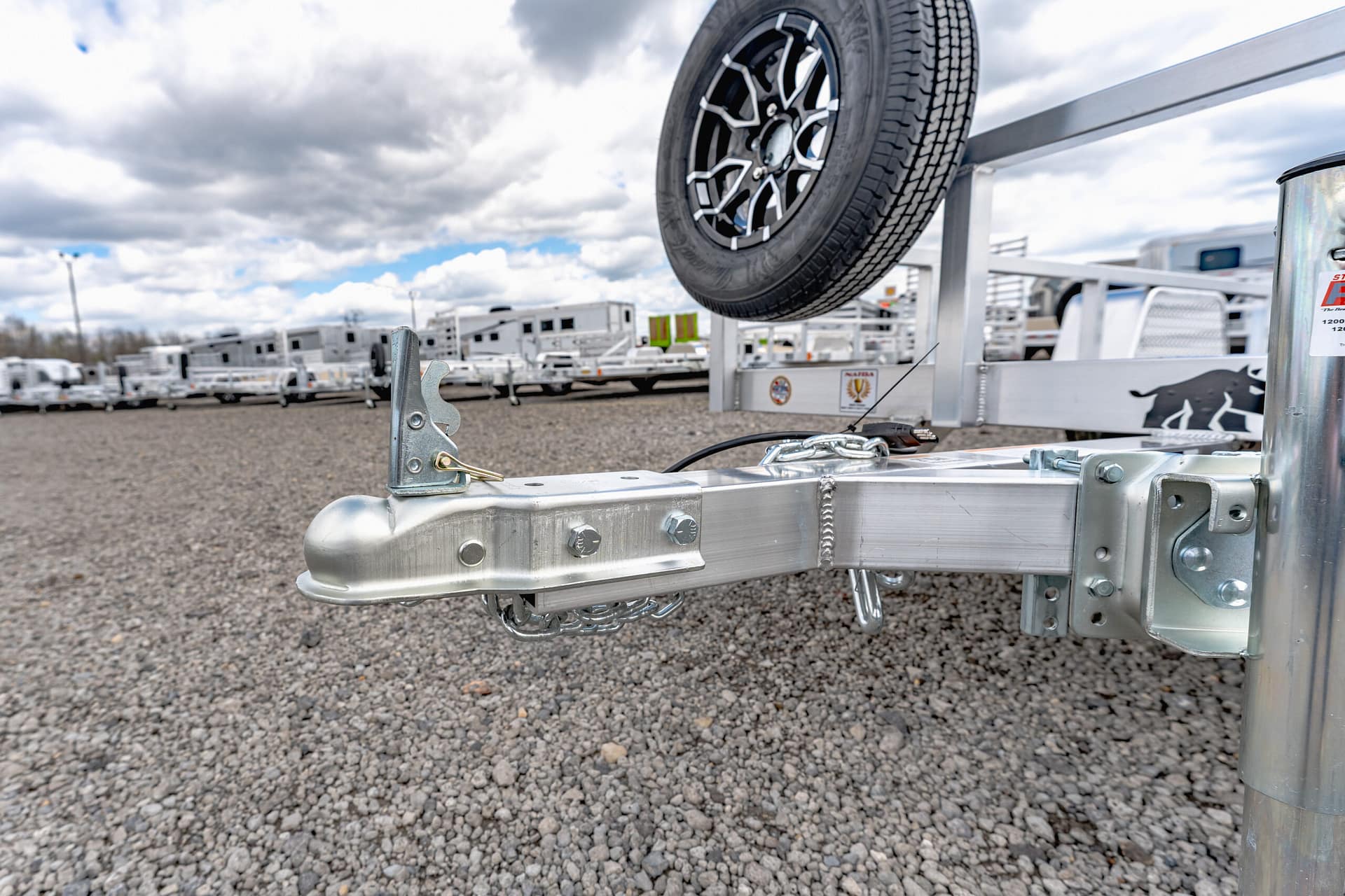 Close-up of a metal trailer hitch with a spare tire mounted above on the trailer frame, in a gravel lot with other trailers in the background.