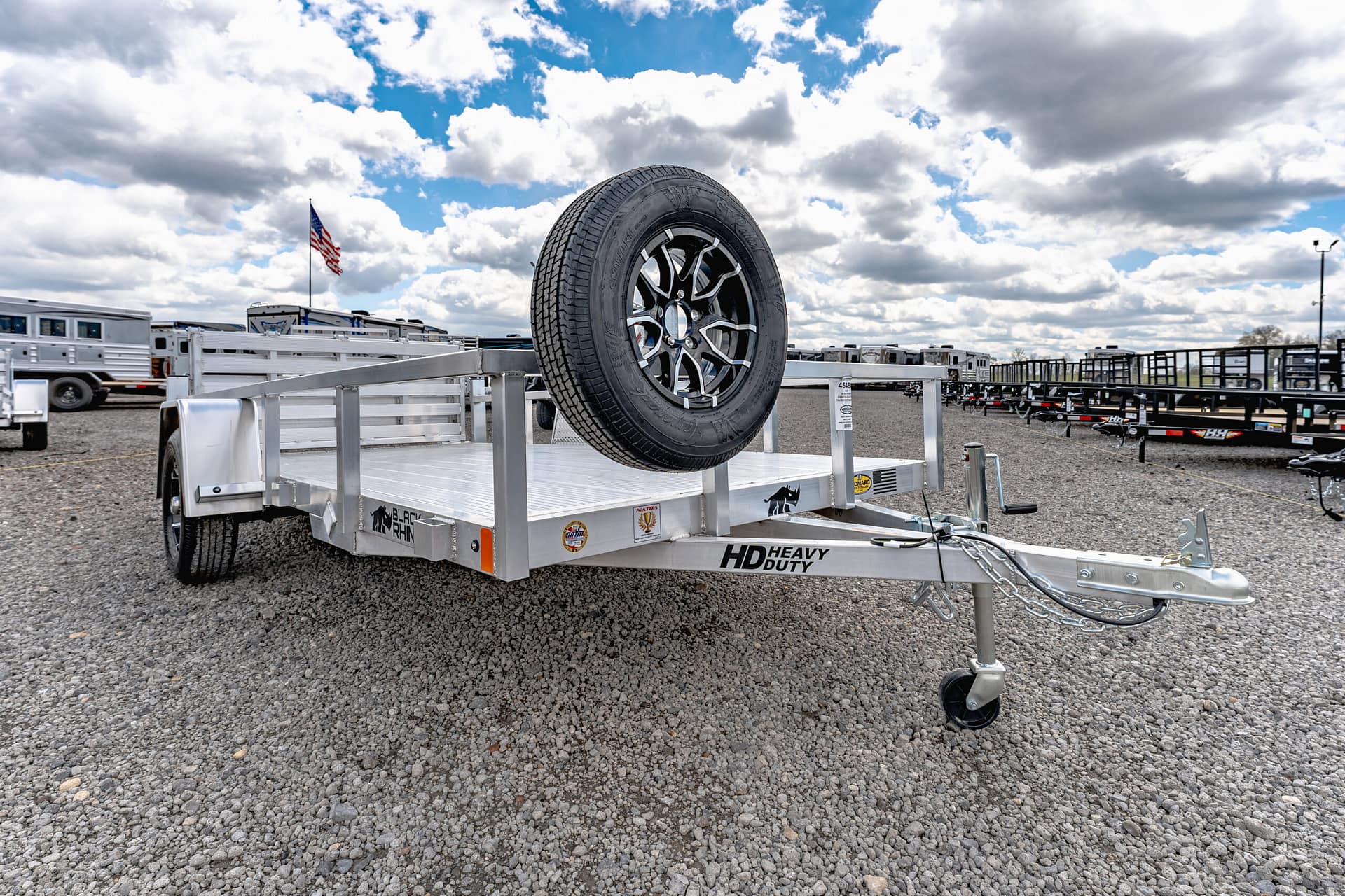 Heavy-duty trailer with a mounted spare tire on a gravel lot, with other trailers and an American flag in the background.