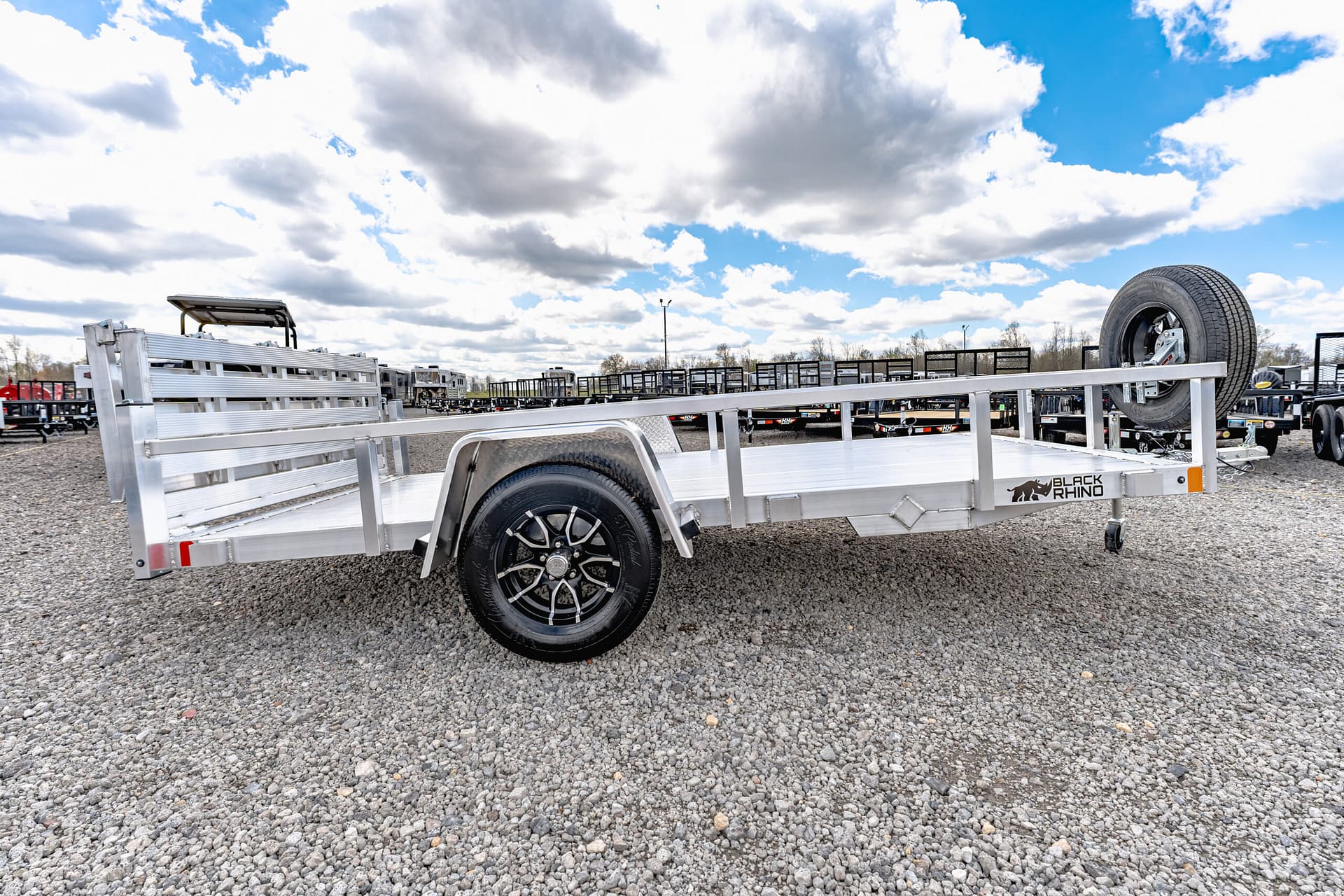 Side view of an aluminum flatbed trailer with a spare tire mounted on the rear rail, in a gravel lot with other trailers in the background under a cloudy sky