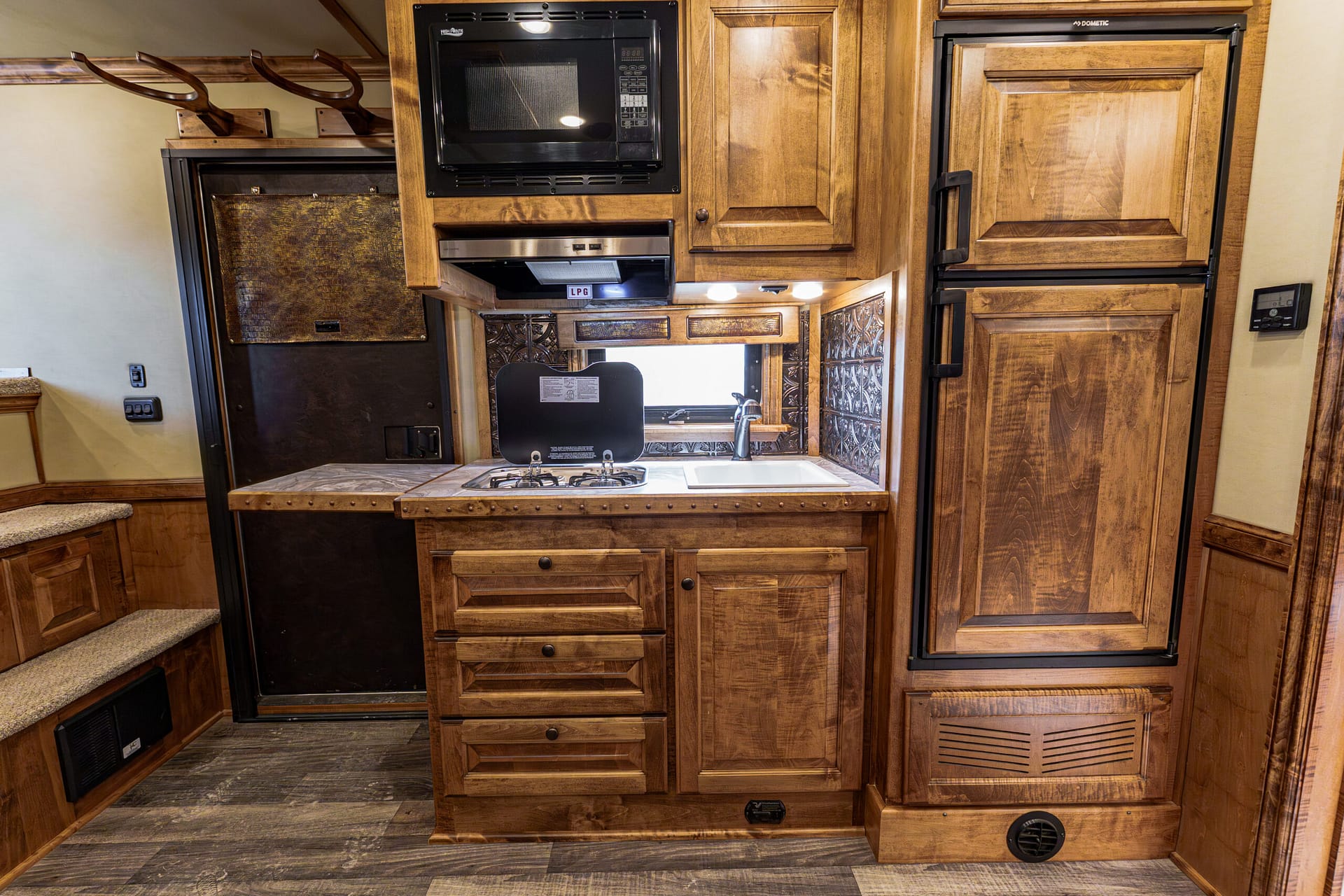 Wood-paneled RV kitchen with a two-burner stove, fold-up cover, microwave above, and wooden cabinets surrounding a sink area.
