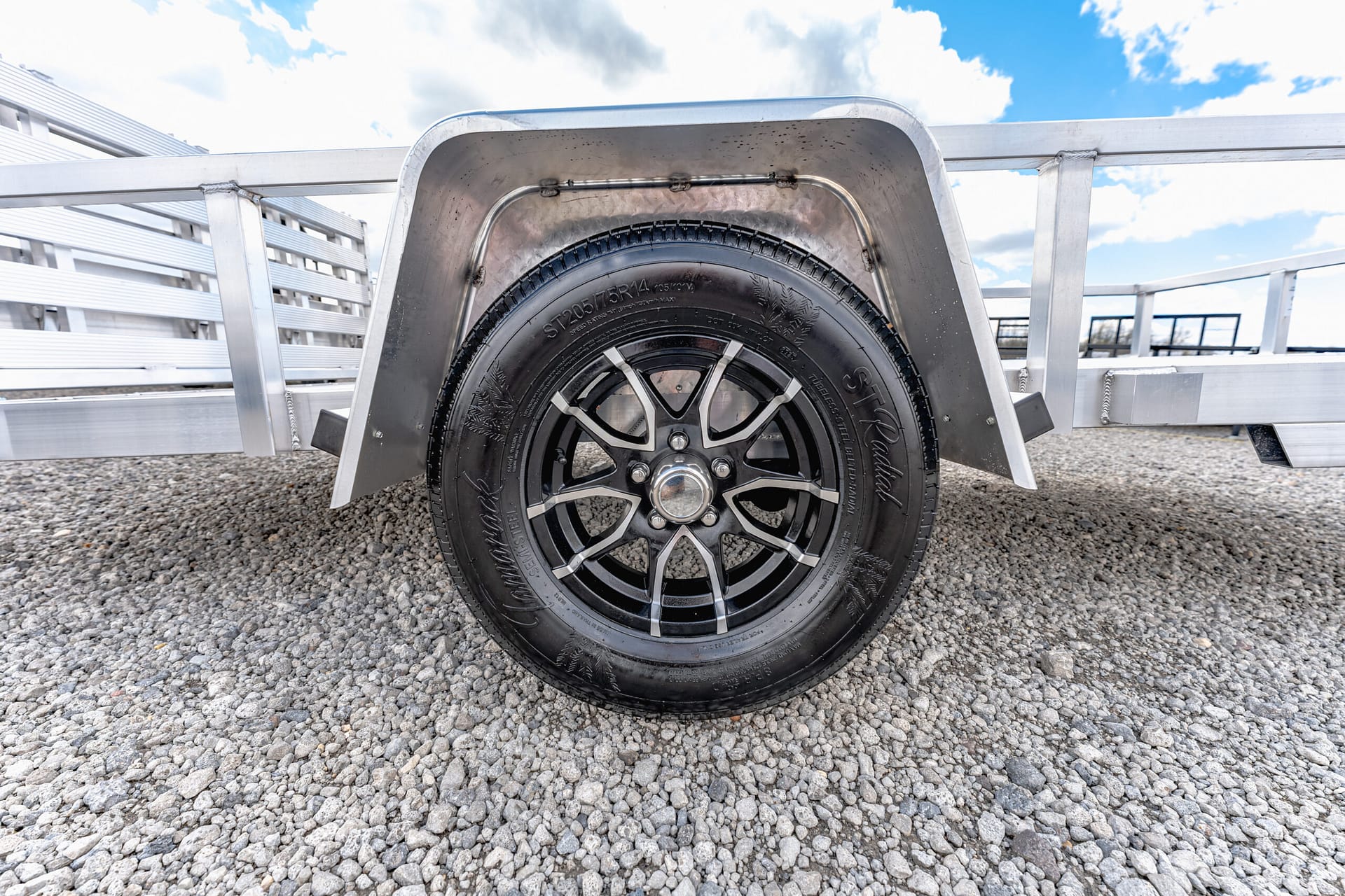 Close-up of a black trailer tire with alloy wheel under a curved metal fender, on a gravel surface with a metal trailer frame nearby.