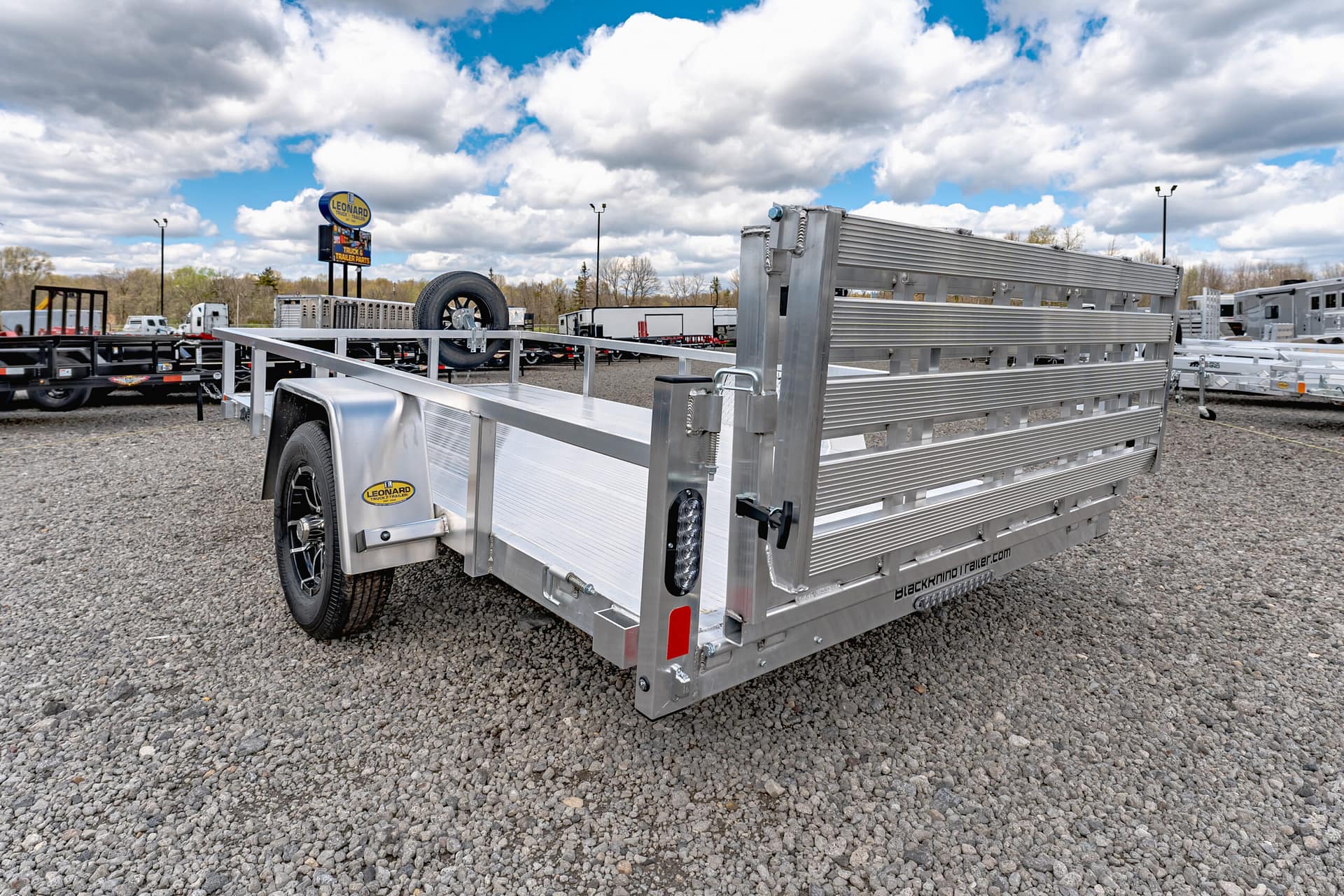 Silver aluminum flatbed trailer with a ramp gate in a gravel trailer lot; Leonard sign visible in the distance.