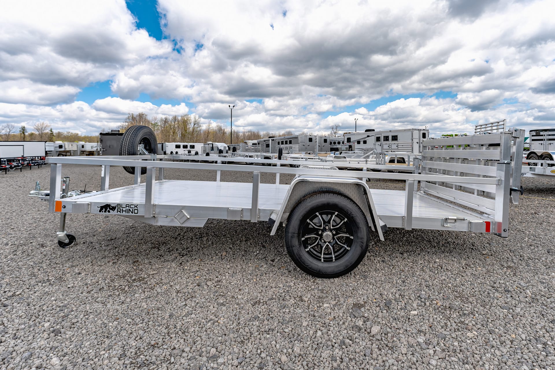Silver utility trailer with Black Rhino logo in a gravel lot, rows of trailers in background under a cloudy sky.