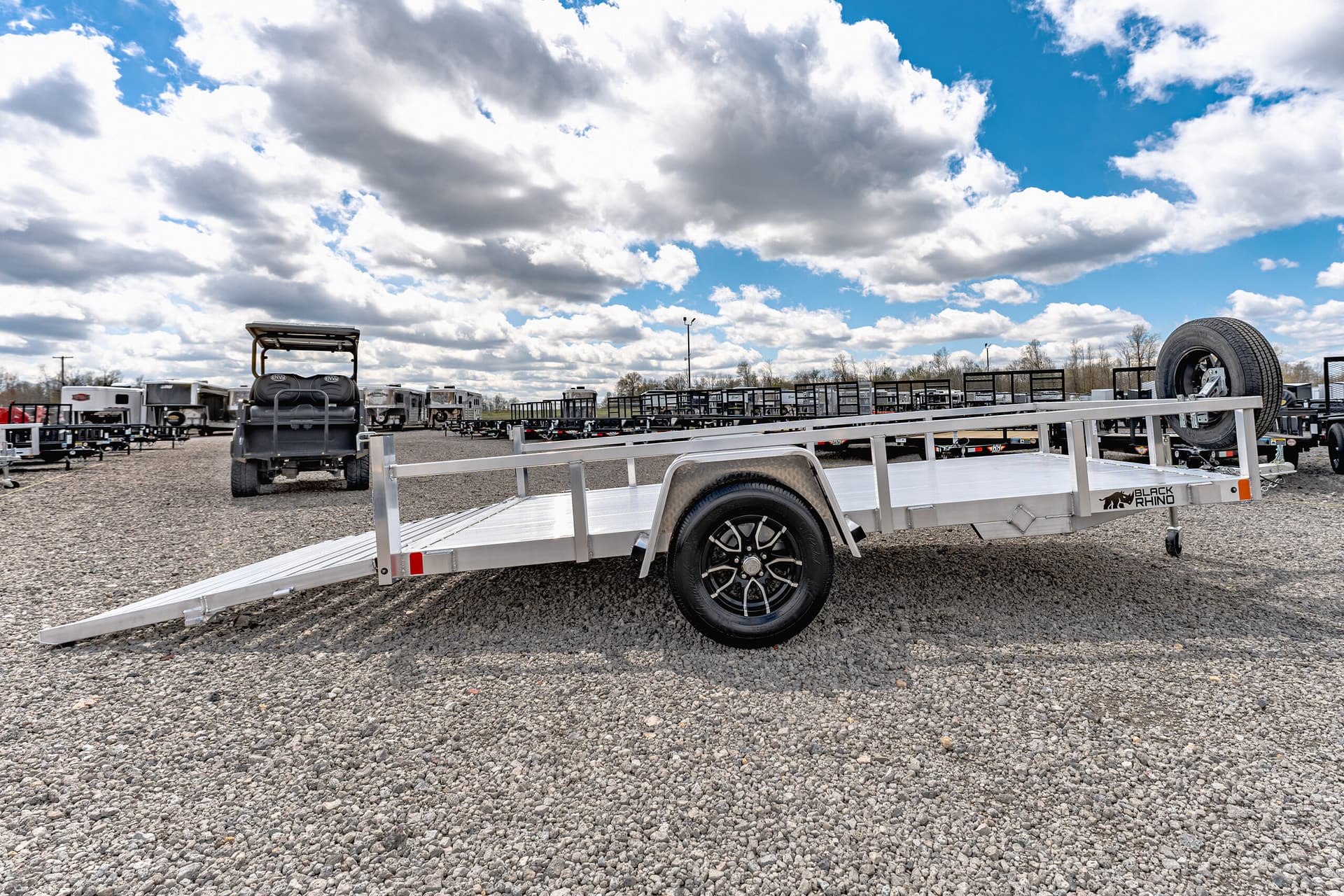 Open lot of flatbed trailers on gravel under a blue sky with fluffy clouds, foreground ramp visible.