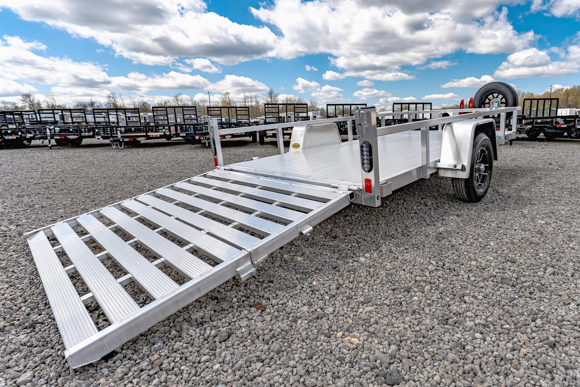 Aluminum truck trailer ramp extended on gravel with rows of trailers in the background and a blue sky with clouds.