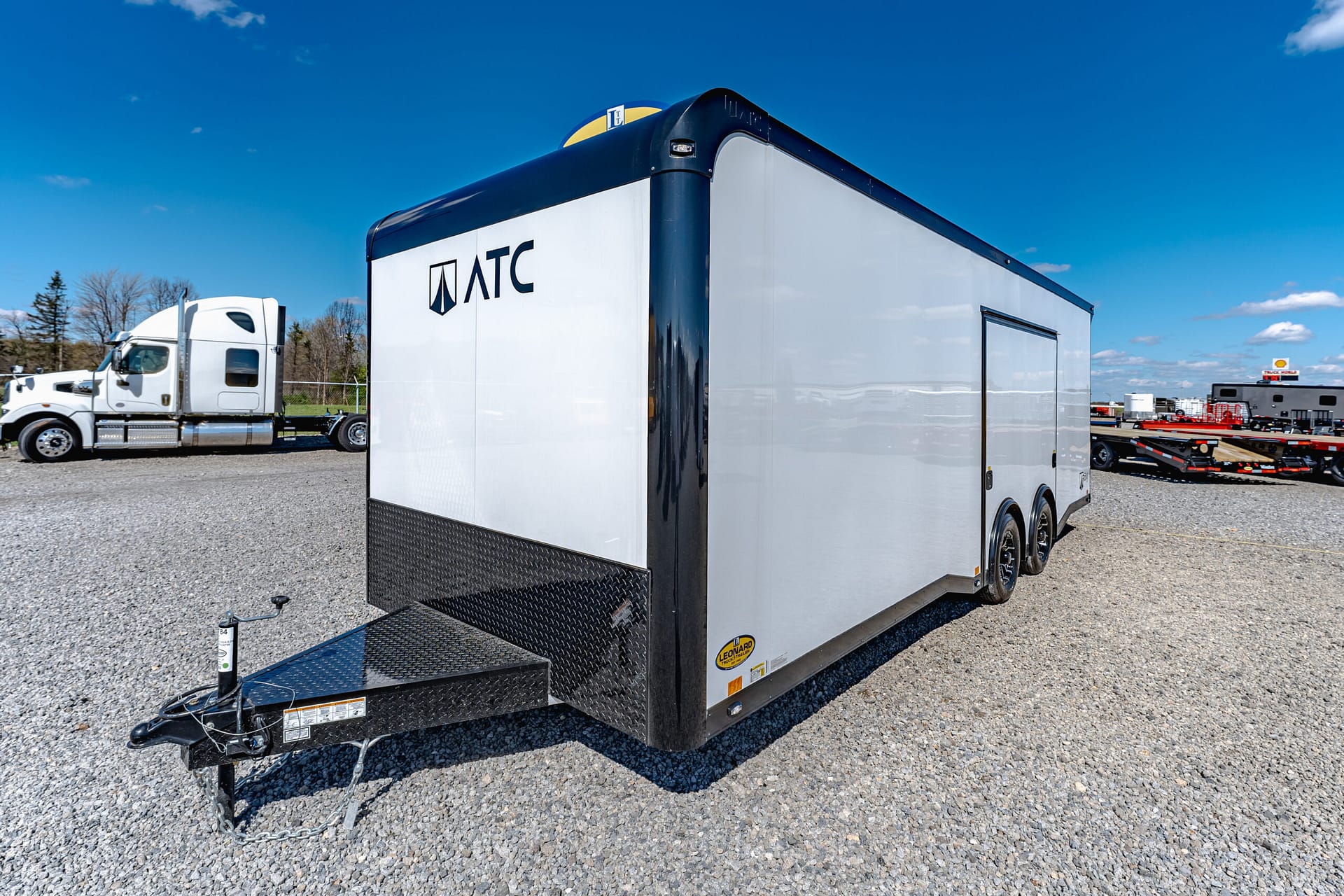 White and gray ATC trailer parked on a gravel lot under a bright blue sky, hitch and chains in the foreground