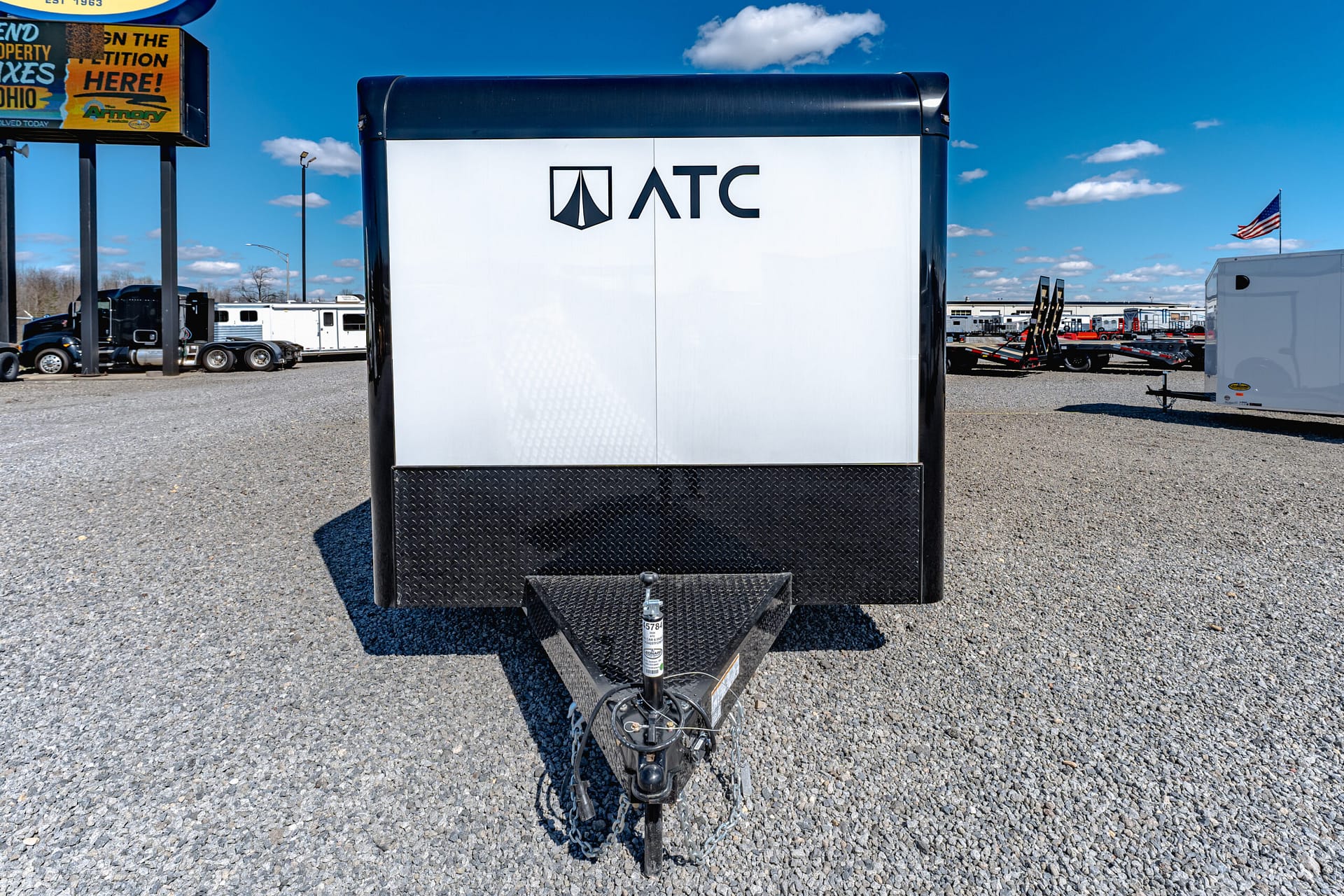 Front view of a white ATC trailer with a black diamond-plate lower front in a gravel lot, blue sky overhead and other trailers in the background.