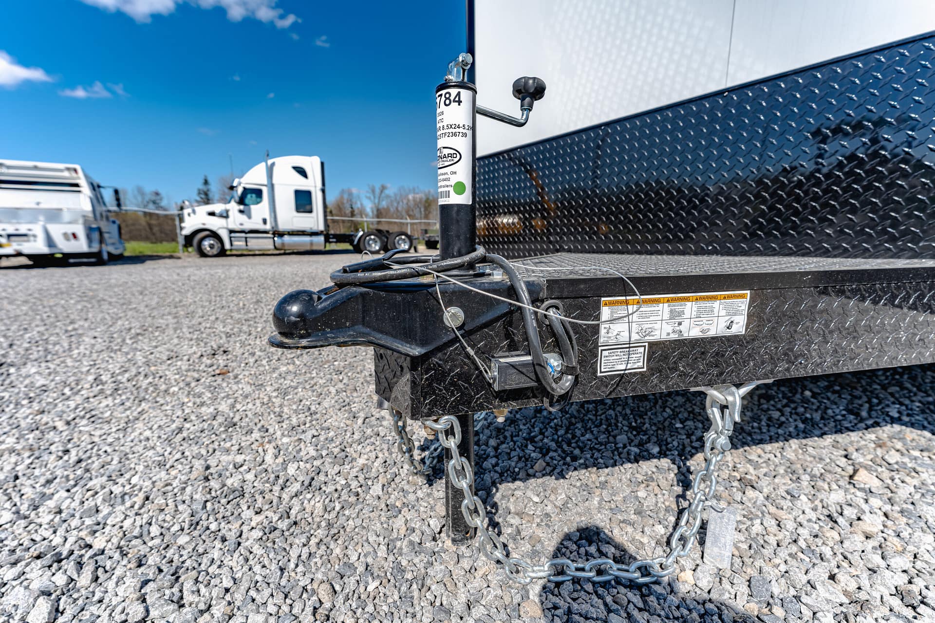 Close-up of a trailer hitch with safety chains and wiring on a diamond-plate trailer, with a gravel lot and trucks in the background.
