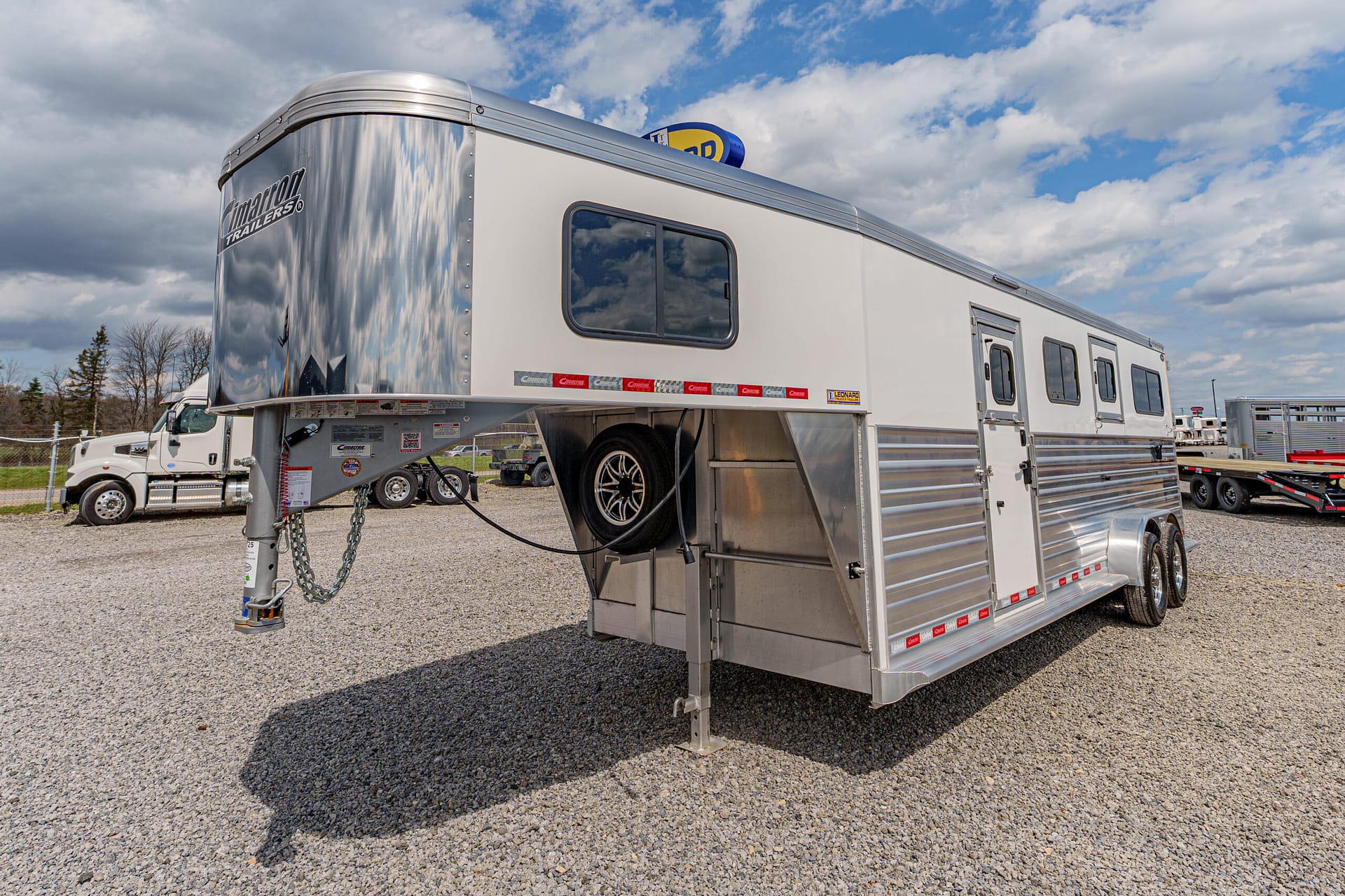 Silver gooseneck horse trailer with a mounted spare tire, parked on a gravel lot.