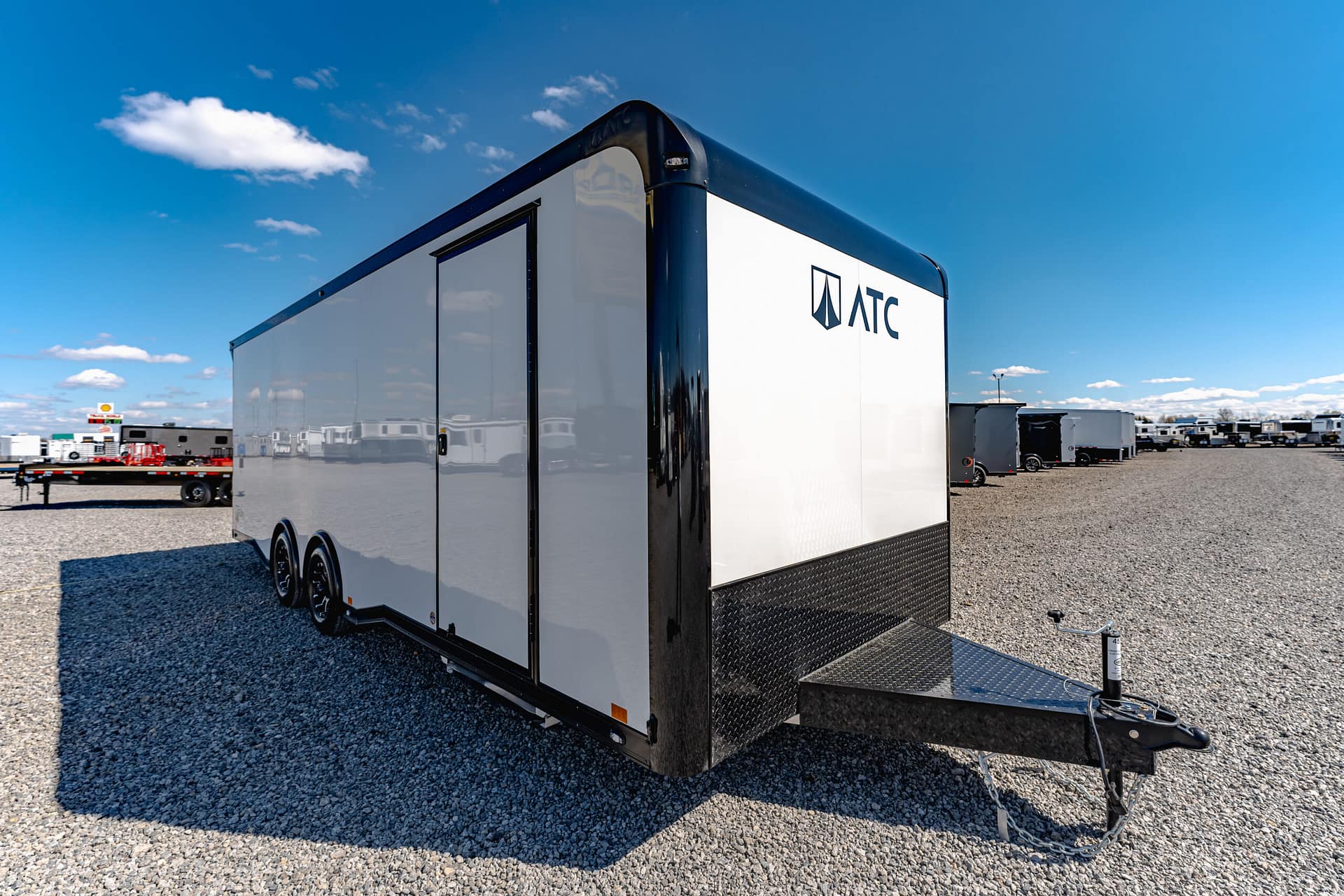 White ATC cargo trailer with black trim visible in a trailer yard on a gravel lot under a clear blue sky, hitch in foreground