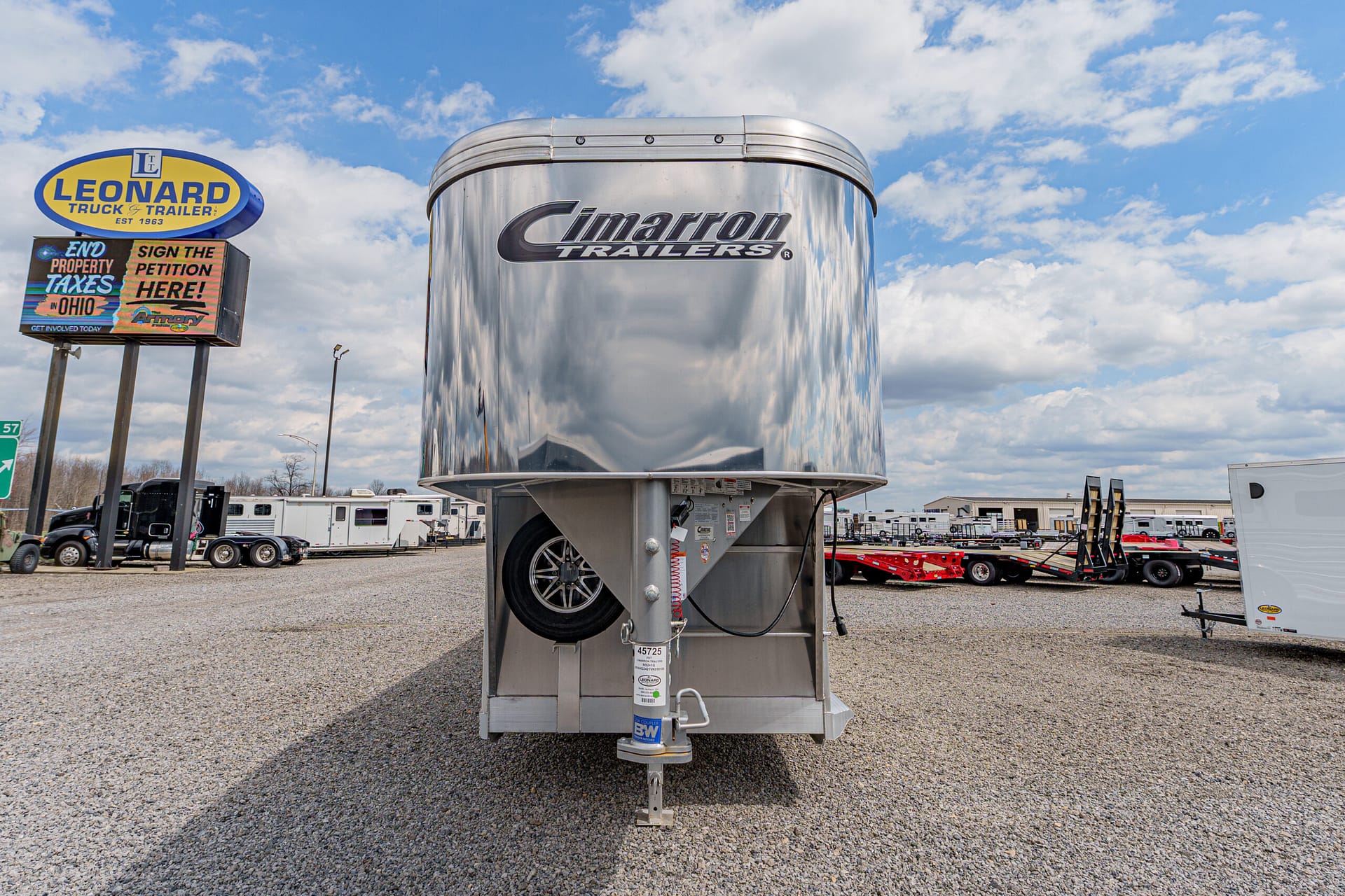 Shiny silver Cimarron Trailers gooseneck trailer on a gravel lot, with a Leonard Truck & Trailer sign in the background.