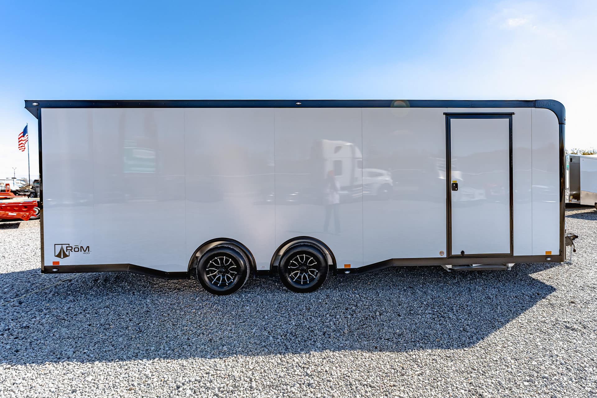 White, enclosed three-axle trailer with a side entry door on a gravel lot beneath a bright blue sky.