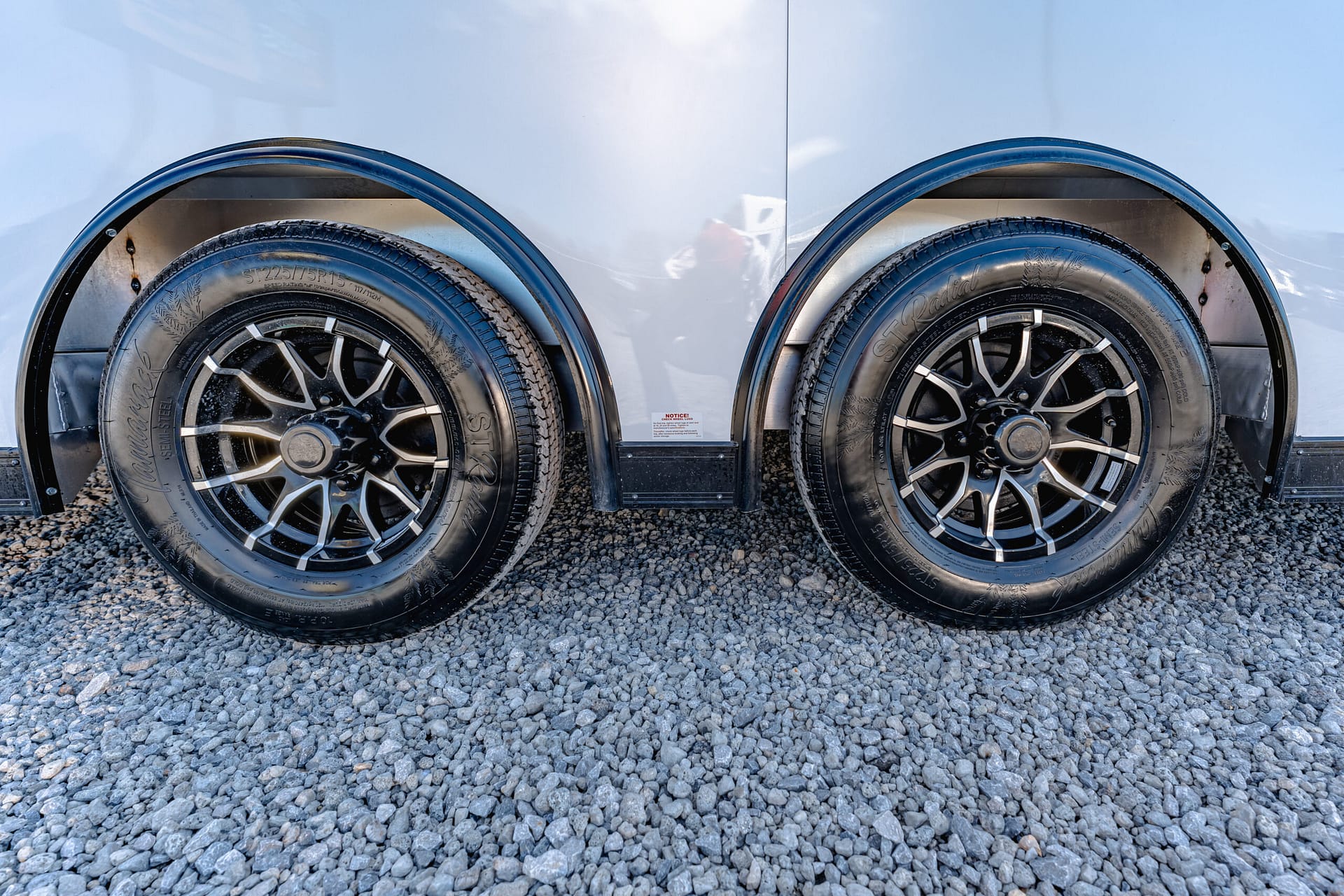 Close-up of two black alloy wheels with tires on a silver trailer, parked on gray gravel.