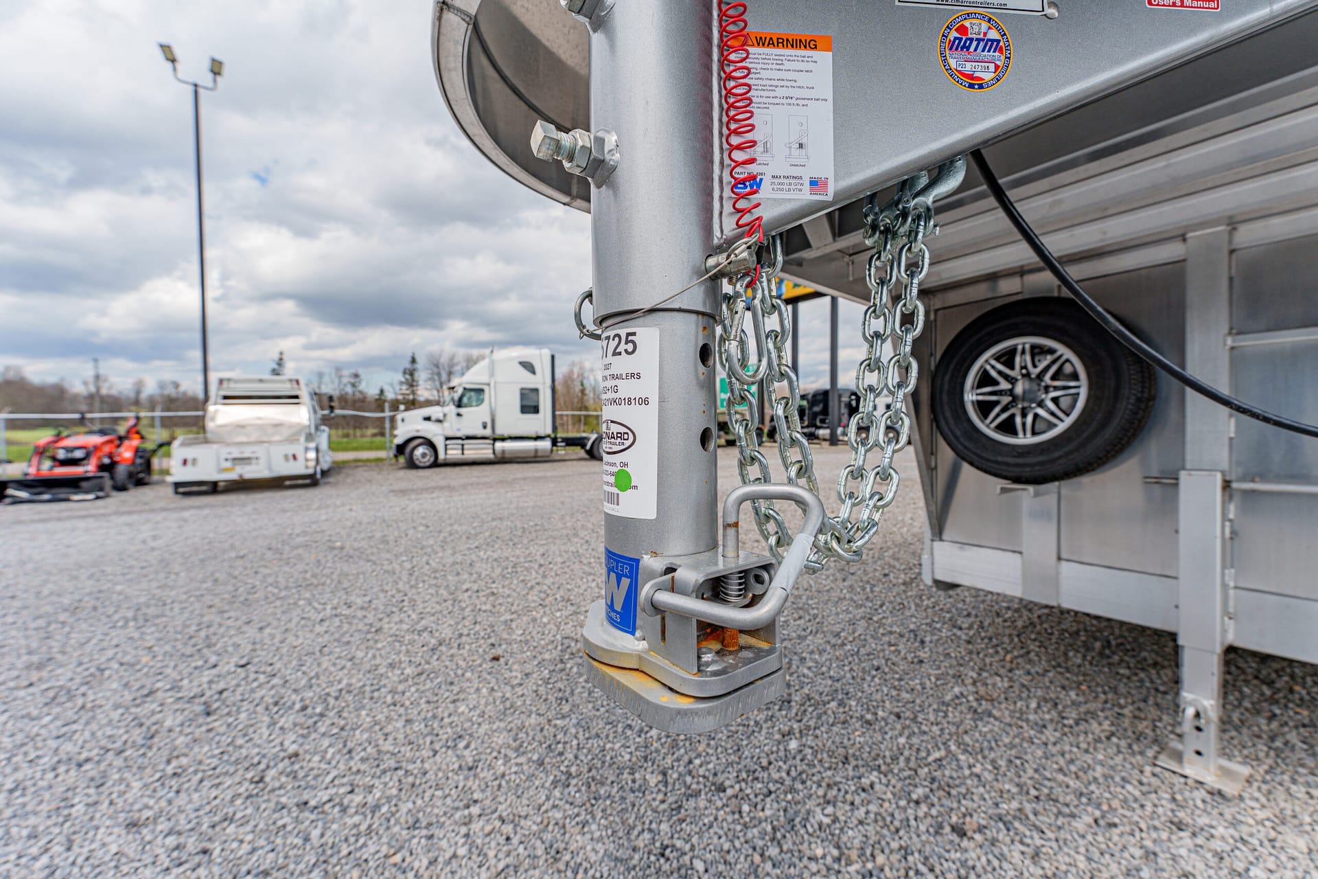 Close-up of a trailer hitch with chains and safety pins on a gravel lot, showing labels and a dual-axle trailer","Close-up of a gooseneck trailer hitch with locking pin and attached chains, on a gravel lot","Trailer hitch area with safety chains, coupler, and warning labels visible on the vertical post","Industrial trailer hitch setup: chains, pins, and decals on the hitch in a parking area","Front of a trailer showing the hitch mechanism, safety chains, and warning stickers on a gray post