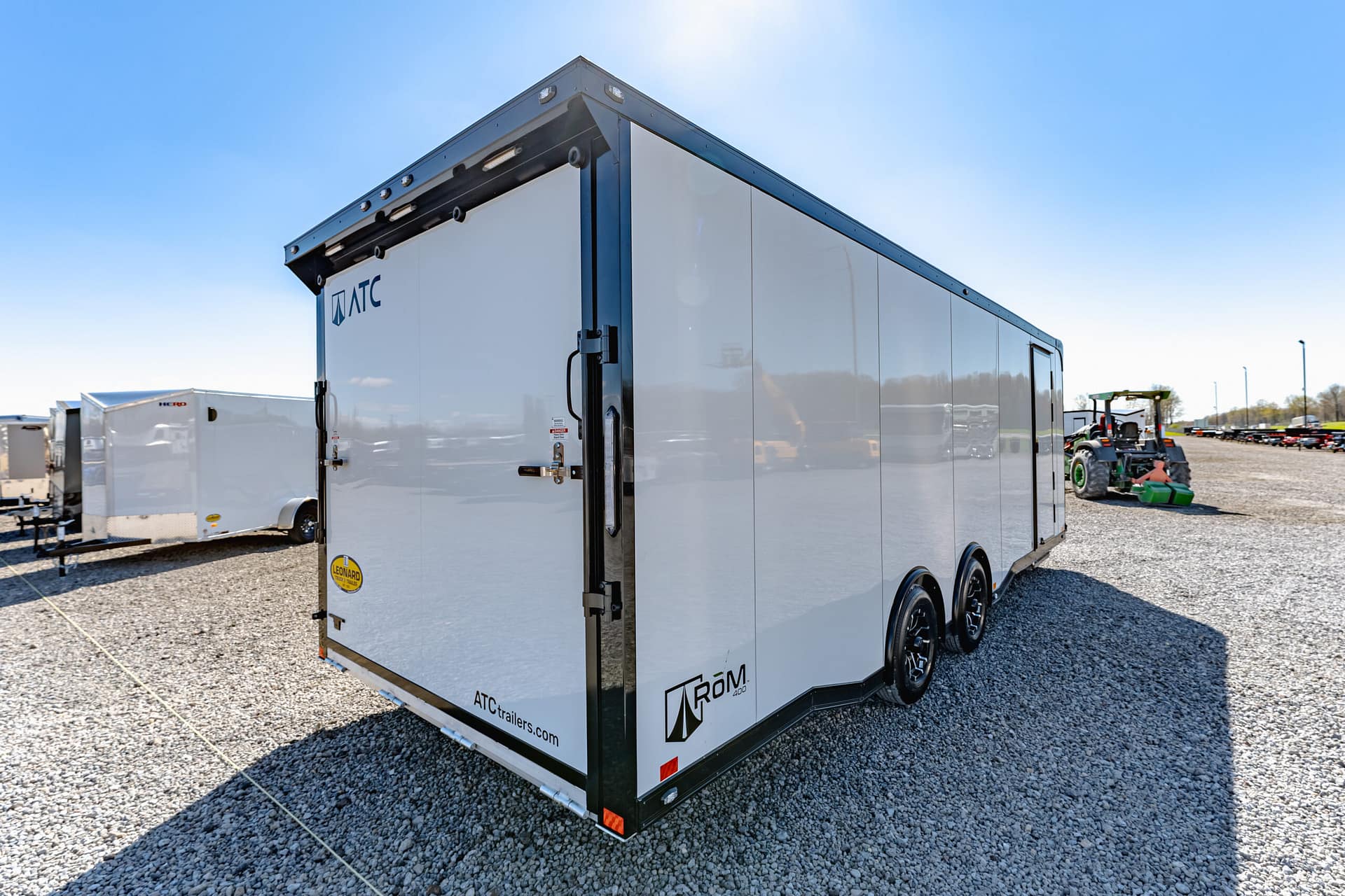 White enclosed trailer with black trim and dual axles, branded with ATC Trailers, parked on a gravel lot under a clear blue sky.