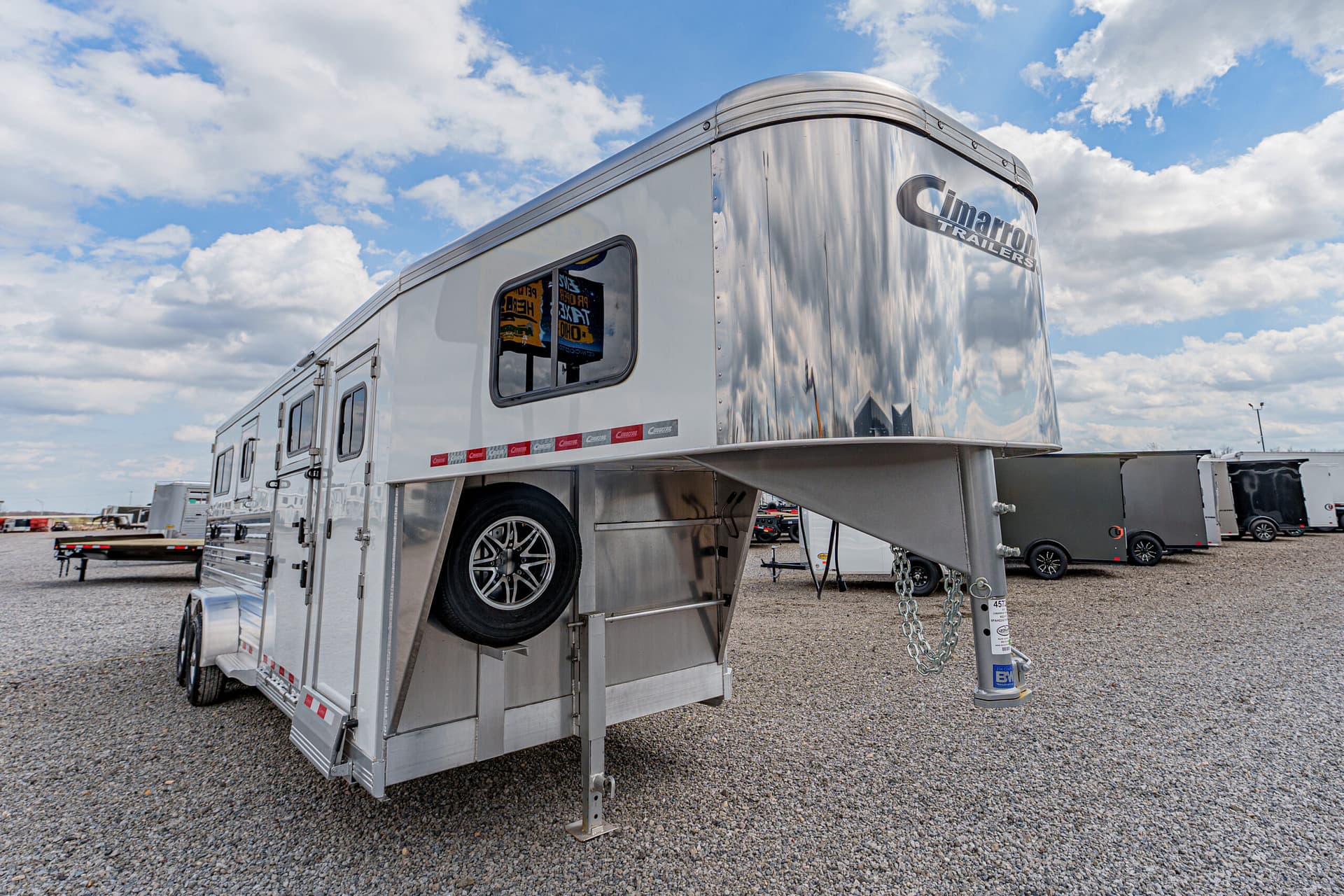 Aluminum gooseneck horse trailer on a gravel lot, spare tire mounted inside the lowered ramp area, Cimarron Trailers logo on front.