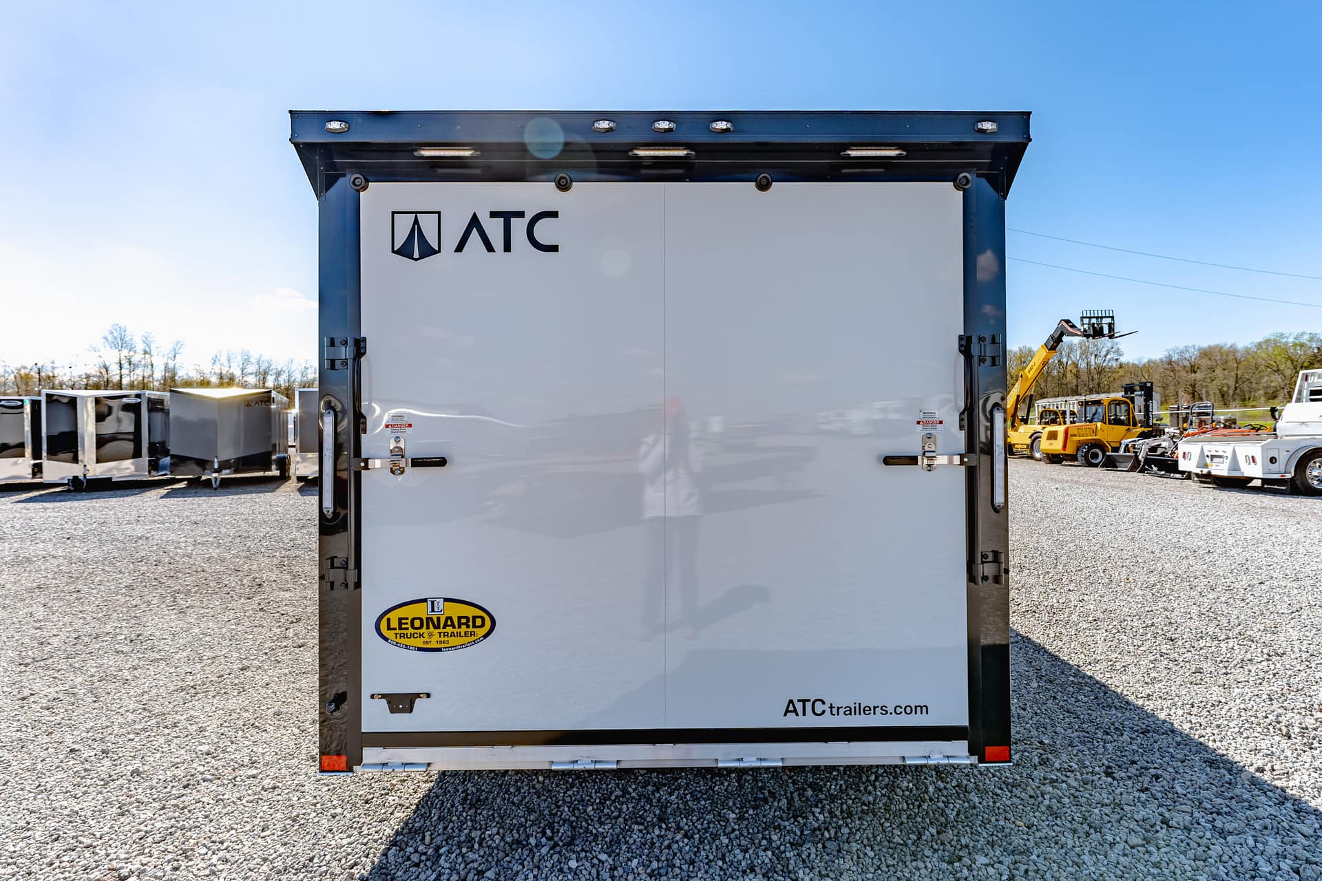 Rear view of a white ATC trailer with Leonard logo, parked on a gravel lot with construction equipment in the background.