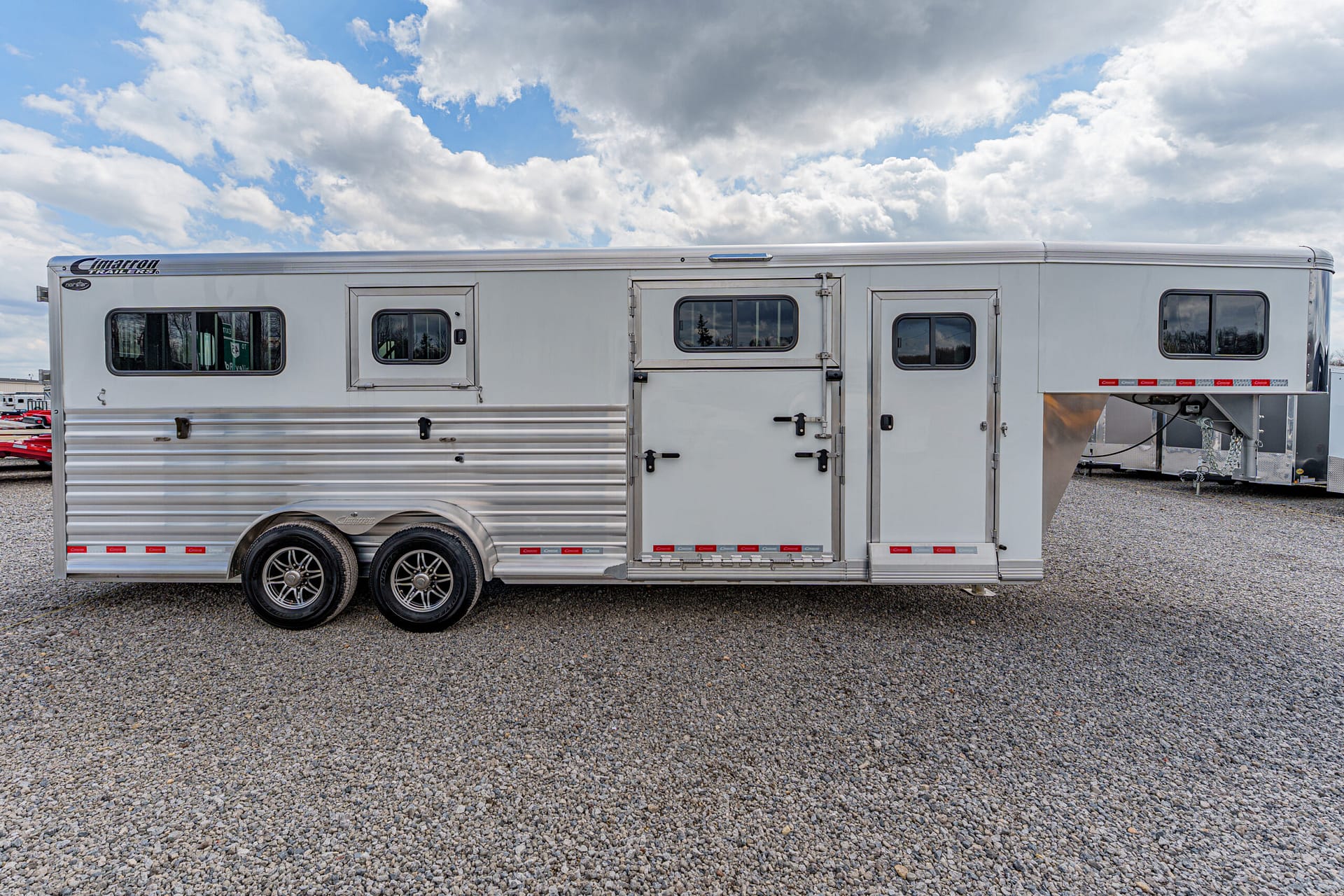 Silver aluminum horse trailer with two axles and multiple windows parked on gravel under a cloudy sky.