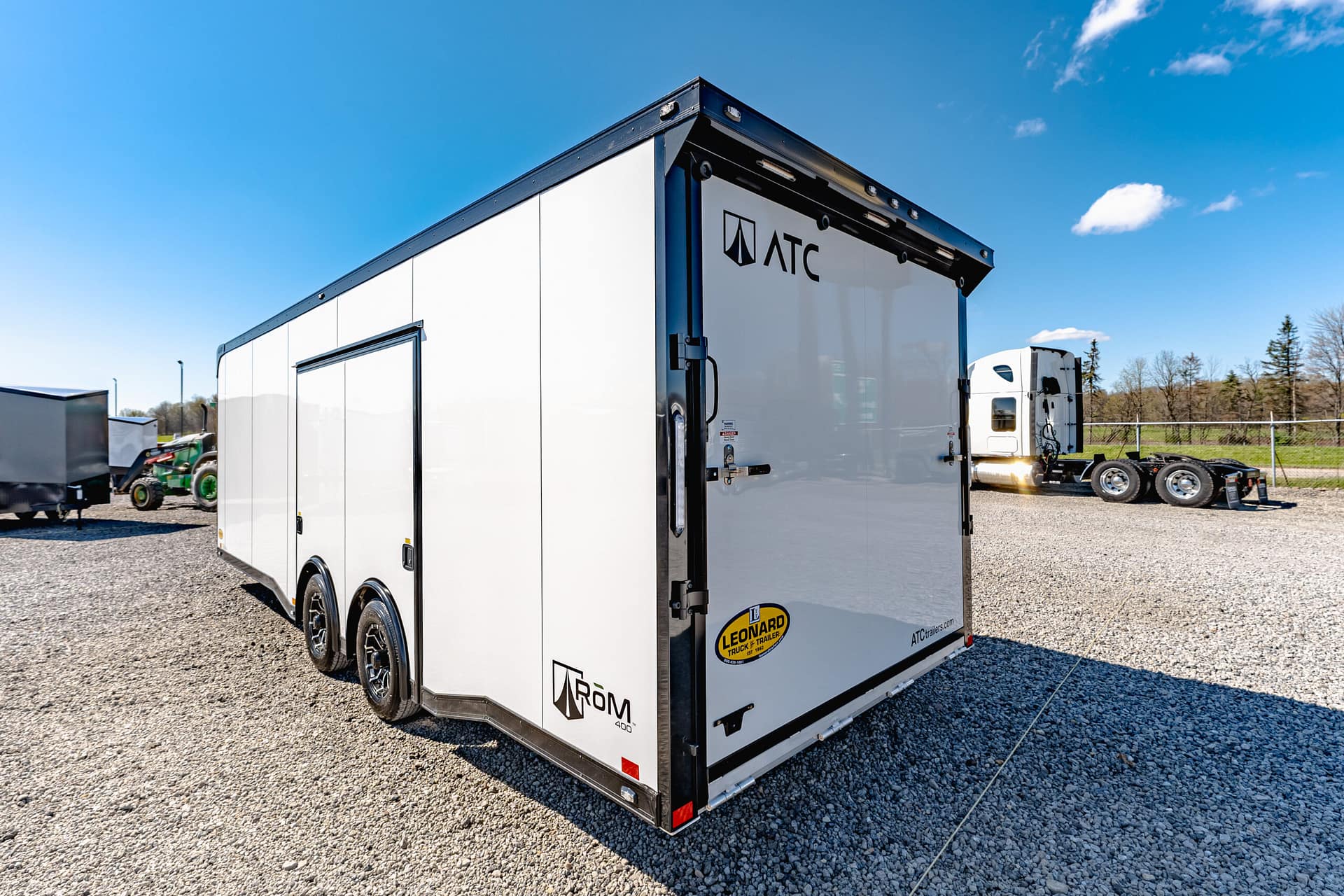 White ATC-branded enclosed trailer parked on a gravel lot under a clear blue sky, with other trailers in the background.
