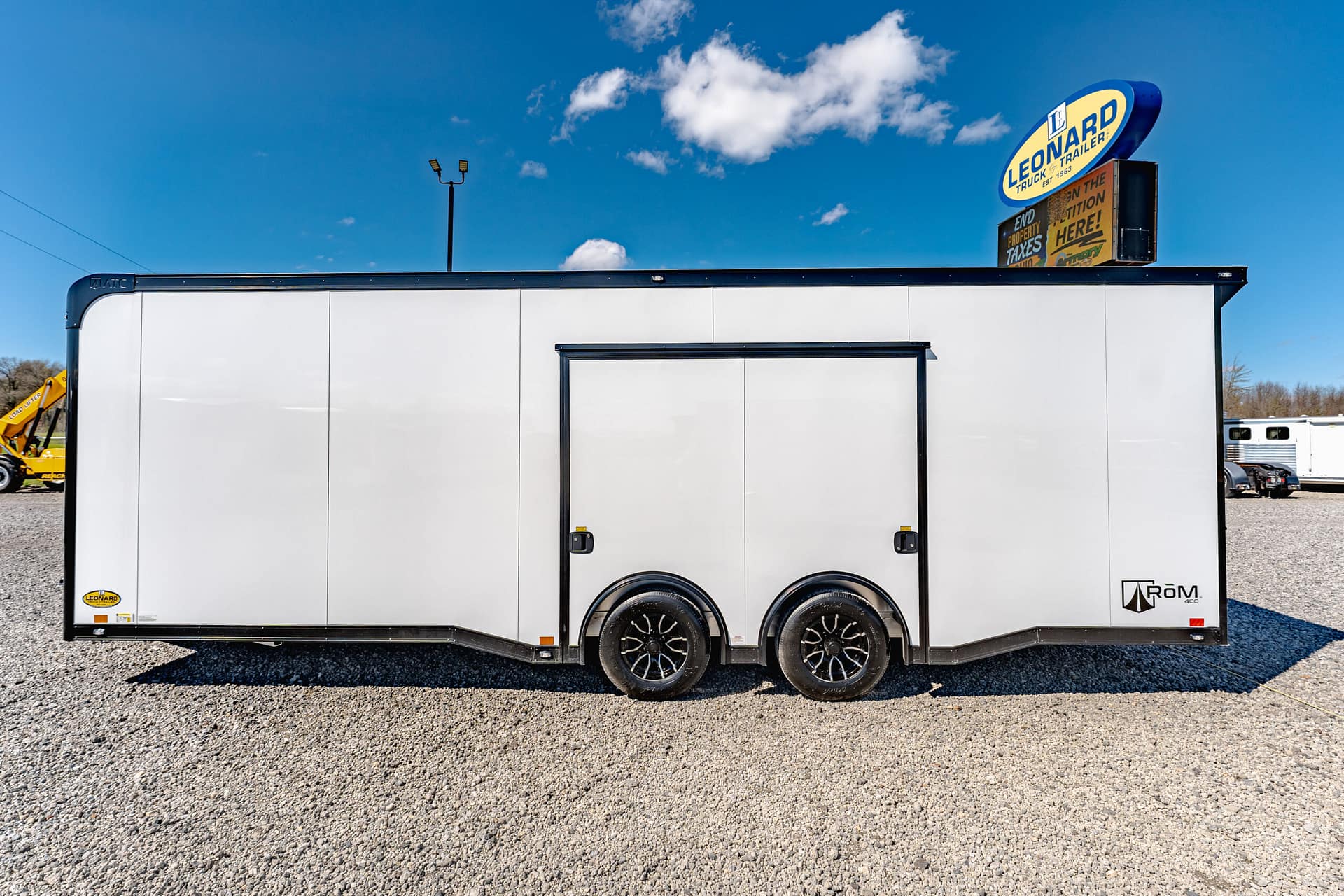 White utility trailer on a gravel lot under a bright blue sky; a Leonard trailer sign is visible above the unit.
