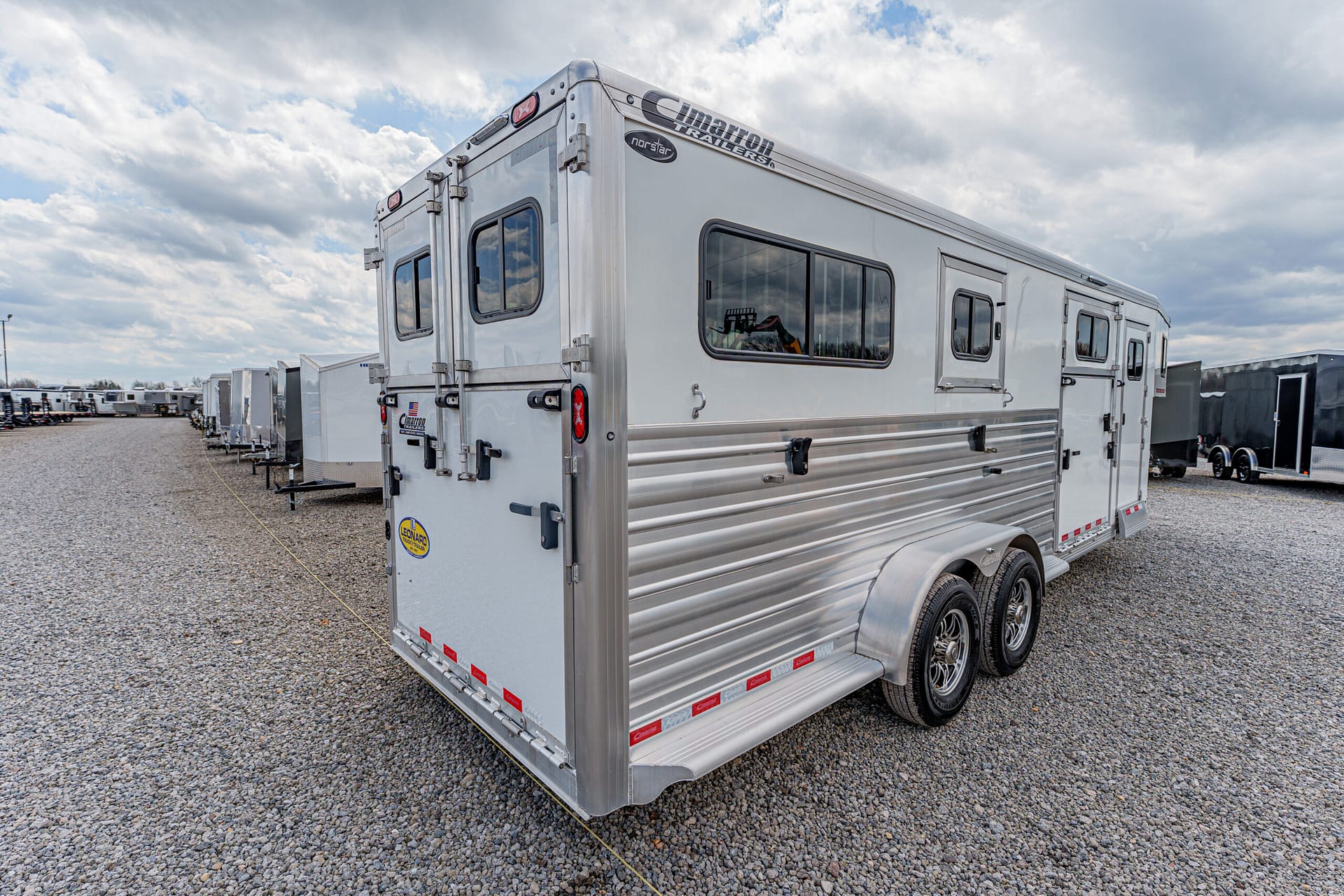 Silver tandem-axle horse trailer with windows, parked on a gravel lot with other trailers in the background.