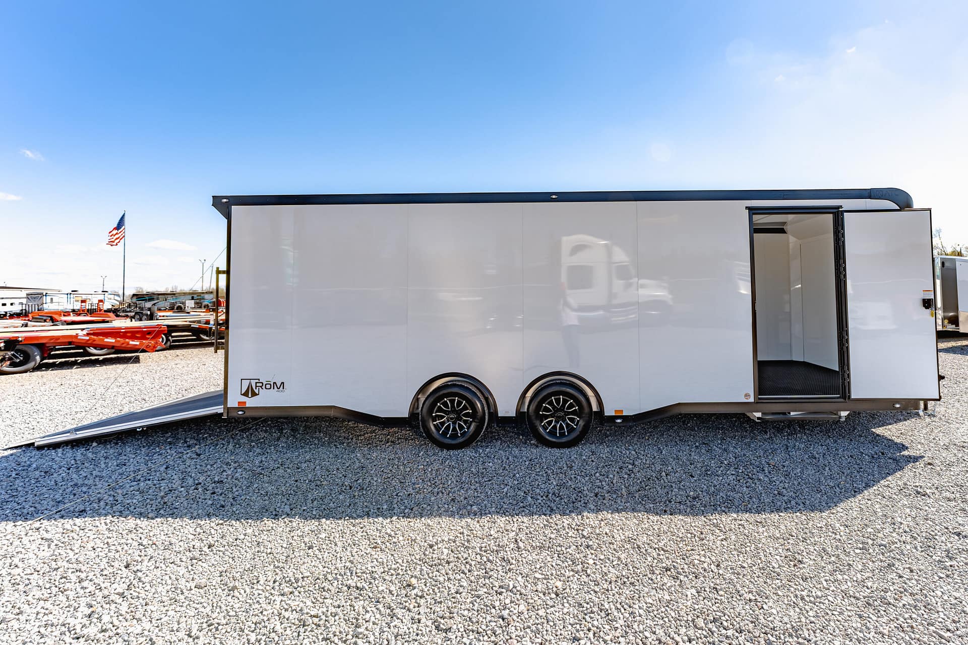 White enclosed trailer with open door and ramp on a gravel lot; American flag and equipment visible in the background under a clear blue sky.