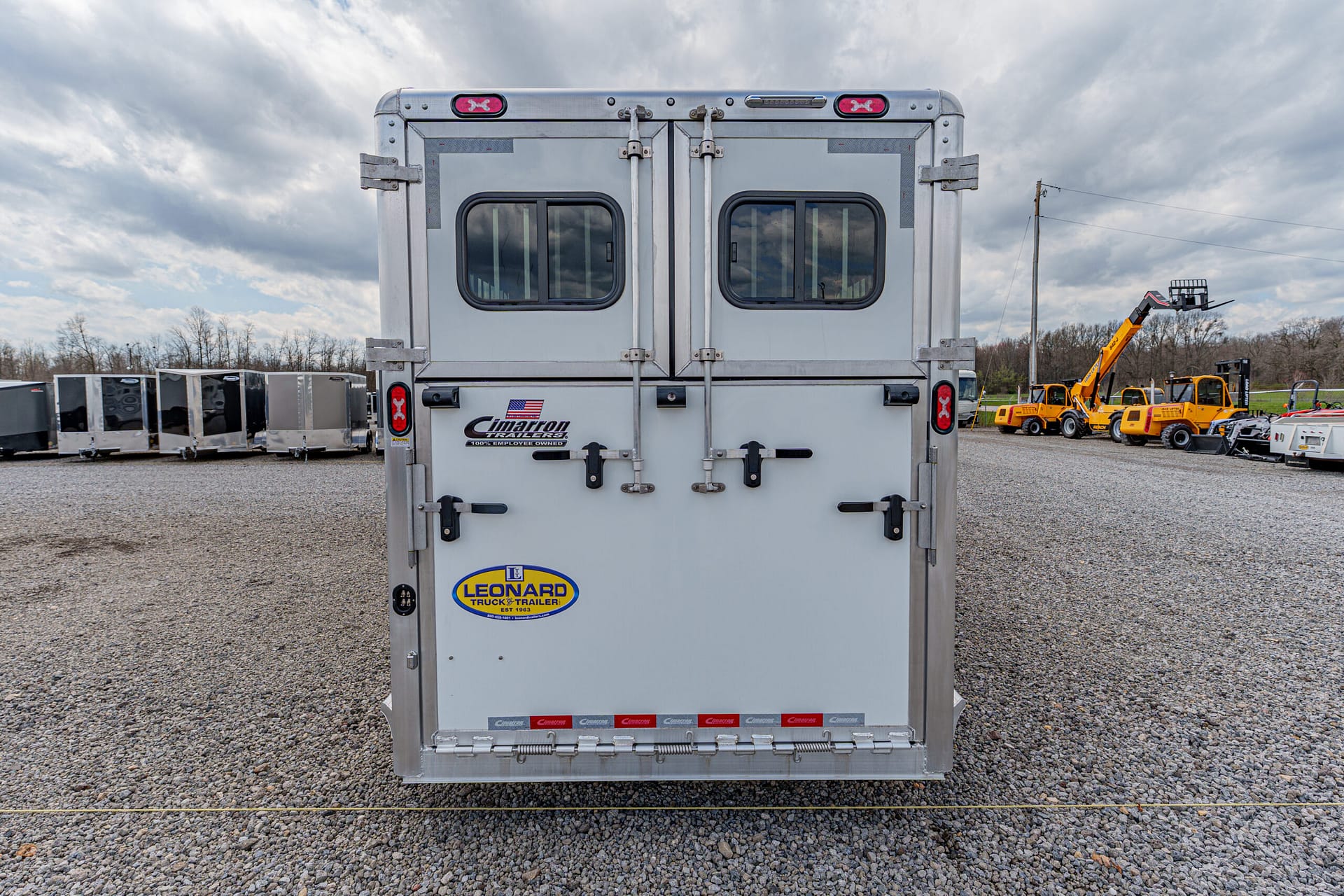 Rear view of a silver cargo trailer with two small windows, double doors, and brand stickers on a gravel lot.