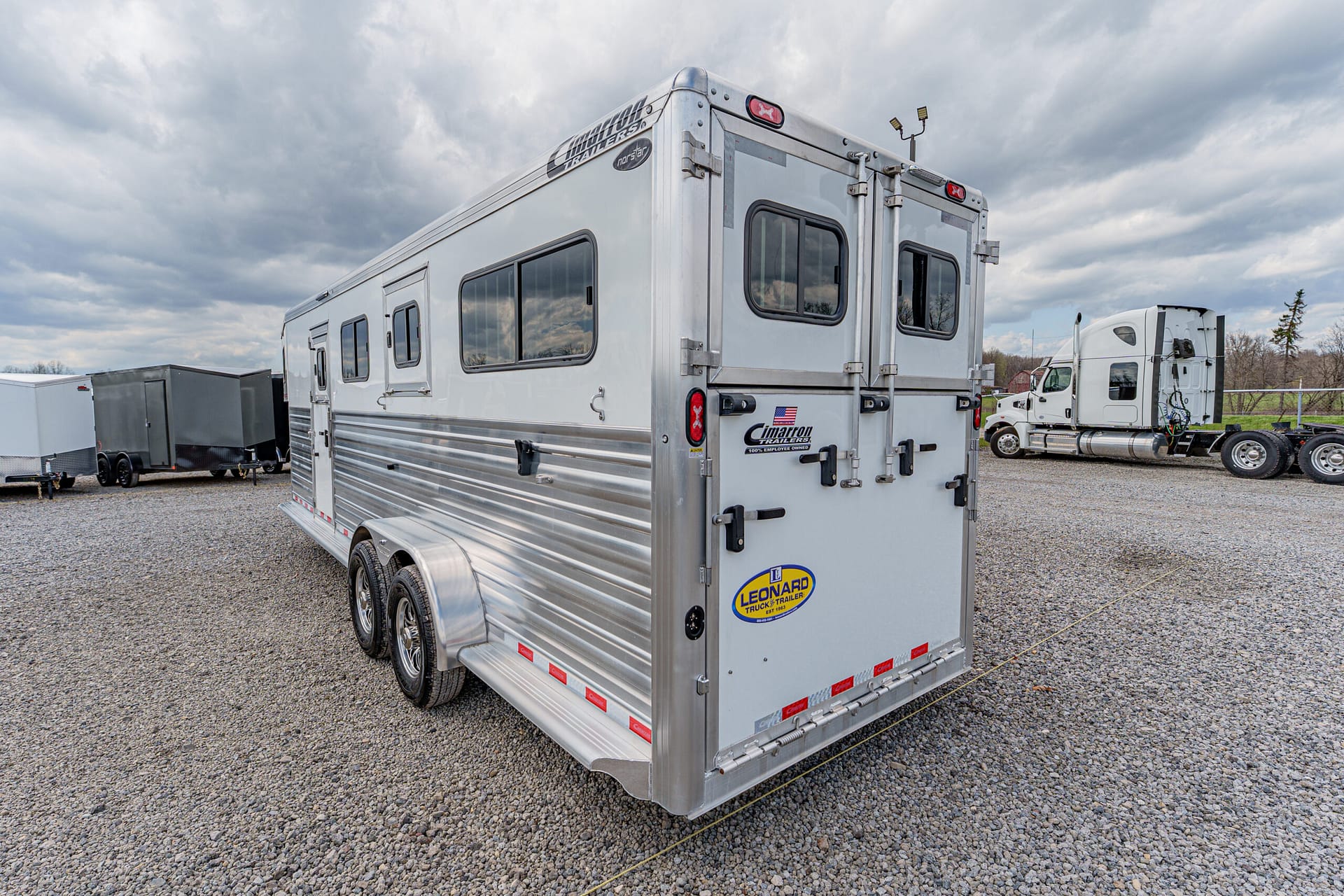 Rear view of a shiny aluminum horse trailer with double doors and two-axle wheels on a gravel lot, with trucks in the background.
