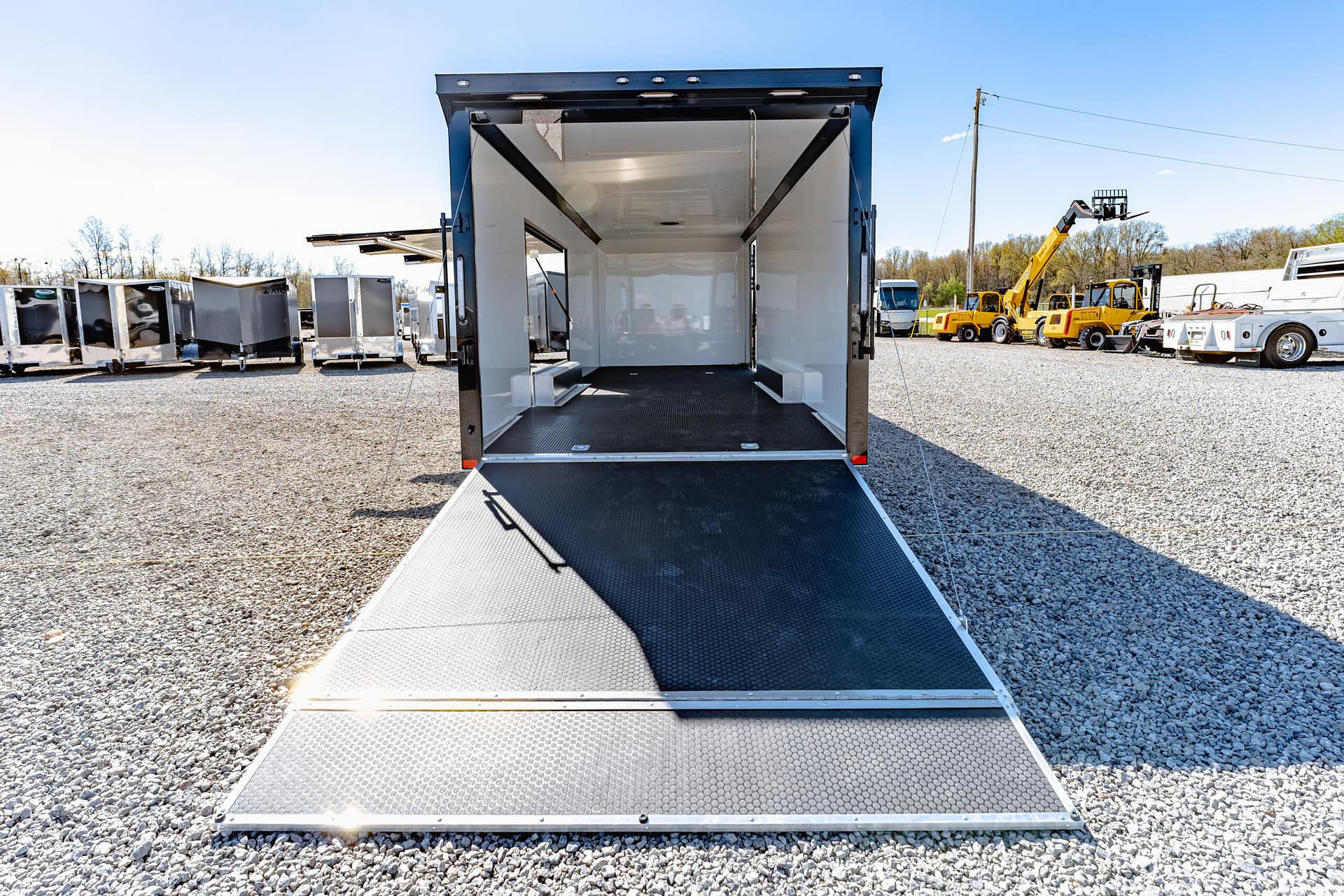 Open cargo trailer with the ramp lowered in a gravel lot, surrounded by other trailers and yellow forklifts.